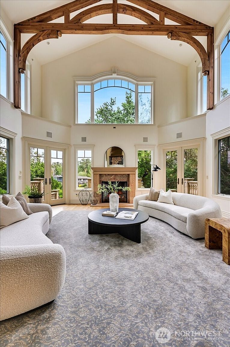 This expansive, high-ceilinged living room features a dramatic exposed timber truss system and a grand stone fireplace as its focal point. The space is elegantly furnished with two curved, cream-colored sofas arranged around a circular black coffee table, all set atop a large, intricate patterned area rug. Large windows and glass doors flood the room with natural light, creating an airy, sophisticated atmosphere perfect for luxury living.
