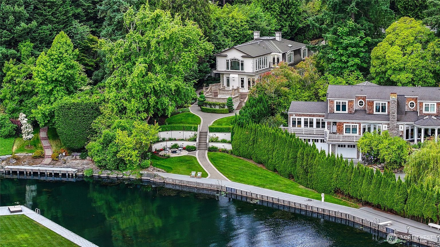 This high-angle aerial view captures a luxurious waterfront estate nestled within a lush, wooded landscape. The property features a multi-level residence with classic architectural details, expansive manicured lawns, terraced gardens, and a private dock extending into the calm water. The scene conveys a sense of serene, high-end privacy and sophisticated outdoor living.