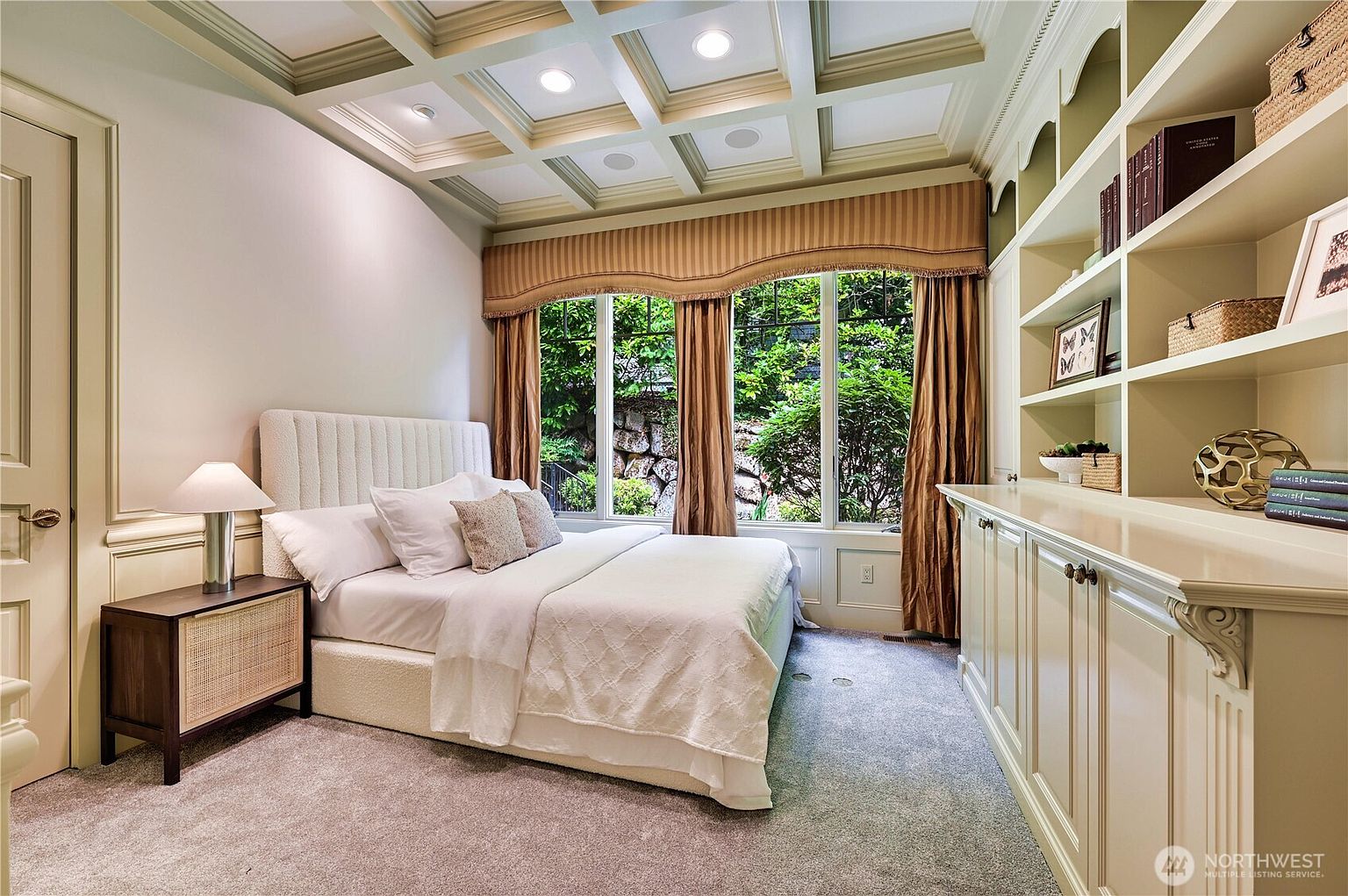 This elegant guest bedroom features a coffered ceiling with recessed lighting and a plush, neutral-toned bed set against a backdrop of large windows with classic drapery. To the right, extensive built-in cabinetry and shelving provide ample storage and display space, creating a sophisticated and organized atmosphere. The room is carpeted and maintains a warm, inviting aesthetic with a view of lush greenery outside.