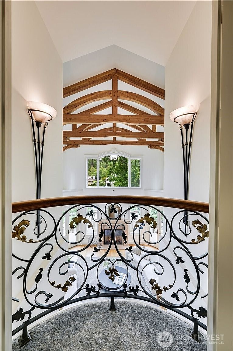 This perspective captures a dramatic view from an upper-level landing, looking down through a decorative wrought-iron railing into a grand, vaulted living space. The architectural design features exposed wooden trusses, elegant wall sconces, and a large arched window that floods the area with natural light. The combination of the ornate metalwork and the soaring ceiling creates a sophisticated, airy, and luxurious atmosphere.