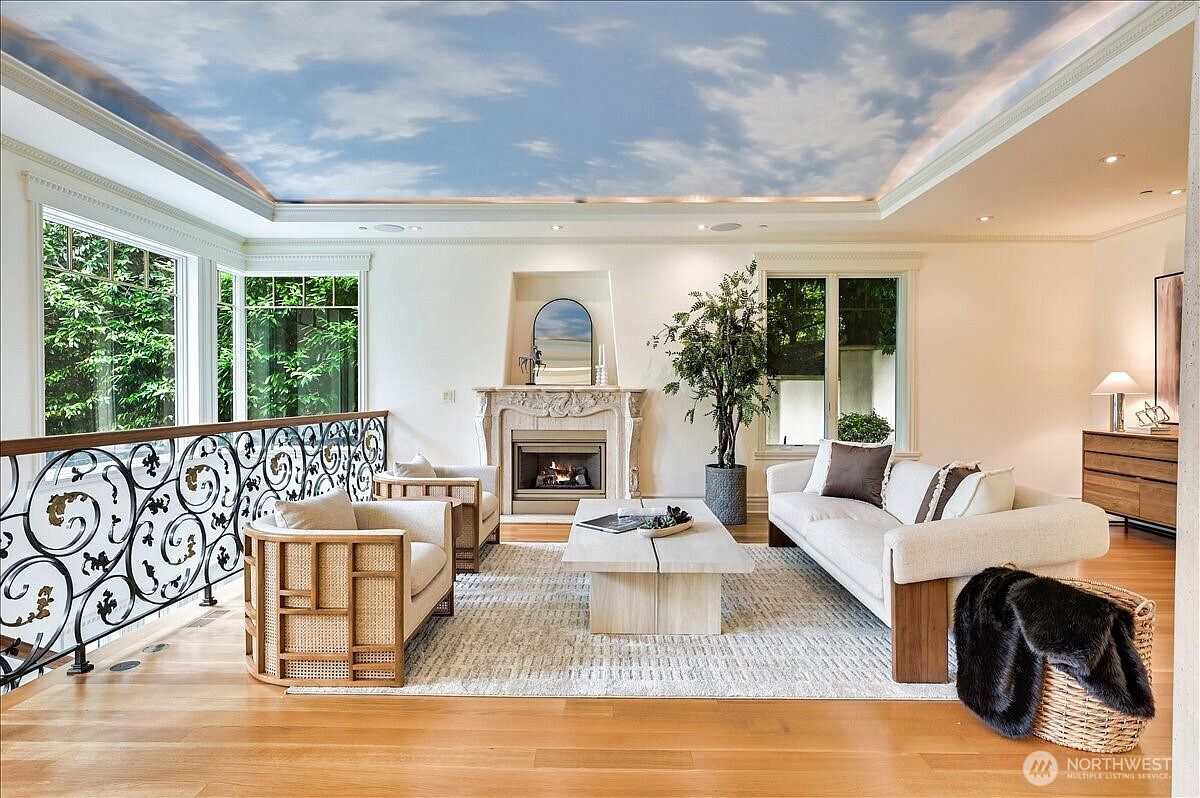 This elegant living room features a striking ceiling mural of a blue sky with clouds, creating an airy and expansive atmosphere. The space is anchored by a stone fireplace, a light-toned area rug, and a mix of modern and cane-accented furniture, all bathed in natural light from large windows. The perspective is a wide, eye-level shot that highlights the sophisticated open-concept layout and the decorative wrought-iron railing overlooking the lower level.