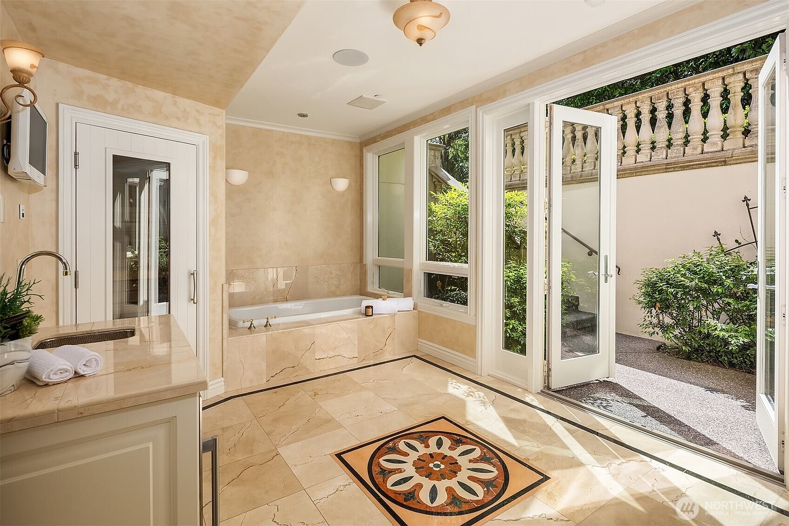 This luxurious primary bathroom features elegant beige marble flooring with a decorative mosaic inlay and matching marble-clad bathtub surround. The space is bathed in natural light from large windows and French doors that open directly onto a private, stone-paved courtyard with a classic balustrade. The room exudes a sophisticated, spa-like atmosphere with high-end finishes and a seamless transition between indoor comfort and outdoor tranquility.