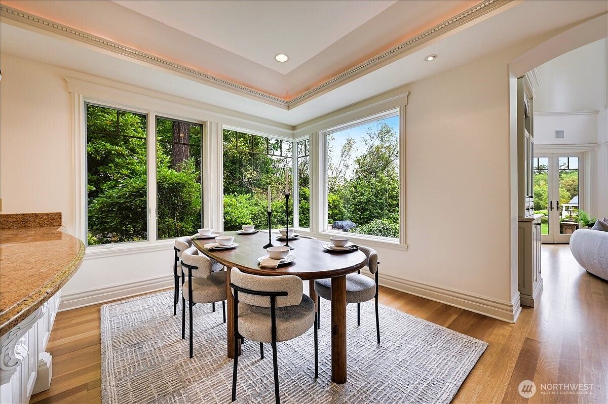 This elegant dining area features a warm, wooden oval table surrounded by modern, upholstered chairs, all set atop a textured area rug. Large, floor-to-ceiling windows wrap around the corner, flooding the space with natural light and offering a serene view of the lush greenery outside. The room is accented by a tray ceiling with recessed lighting and flows seamlessly into an adjacent living space, creating an inviting and sophisticated atmosphere.