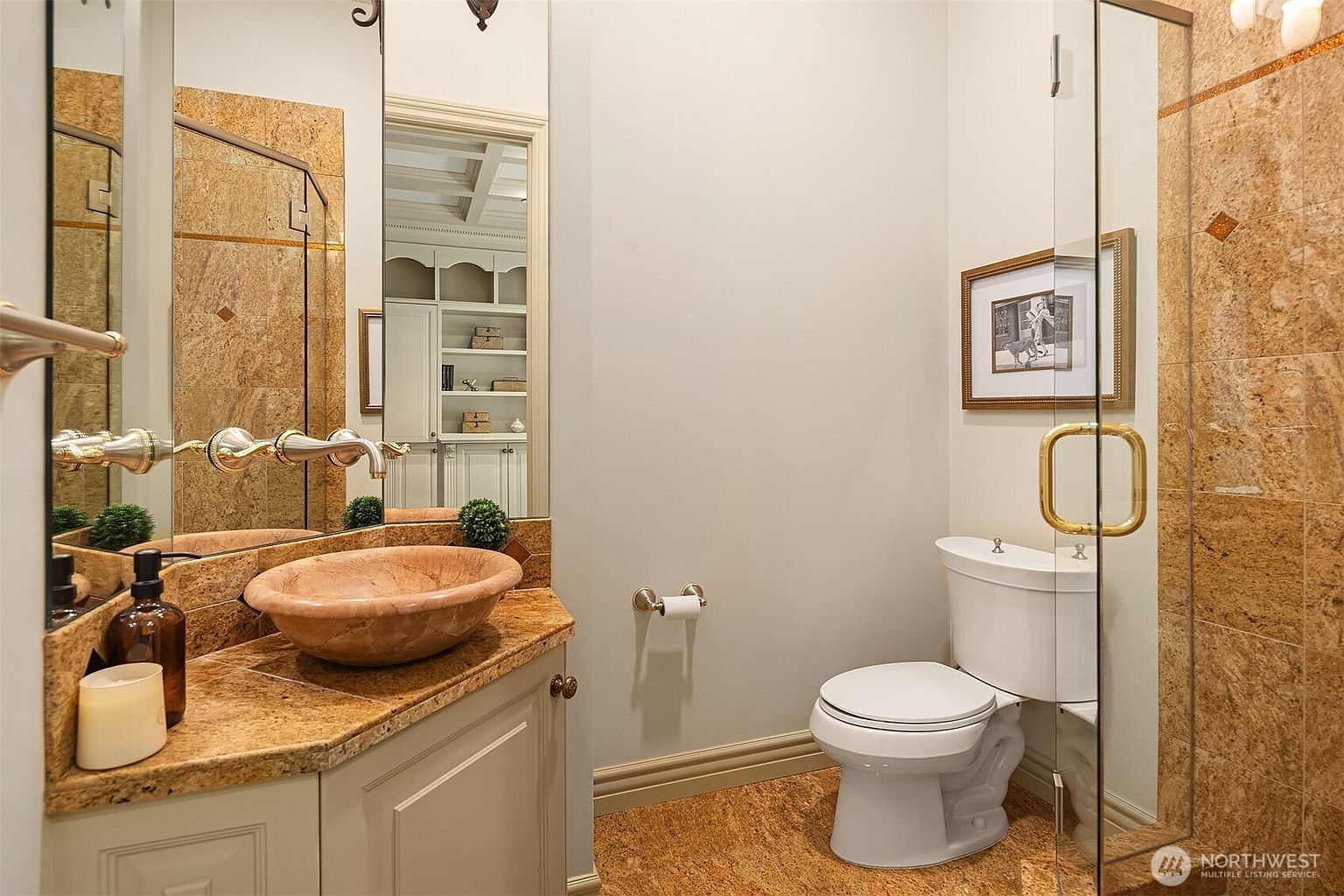 This elegant powder room features a unique stone vessel sink set atop a granite vanity with gold-toned fixtures. The space is accented by warm, textured stone wall tiles and a large mirror that reflects an adjacent room, creating a sense of depth. A classic white toilet and a framed piece of art complete this sophisticated and compact bathroom design.