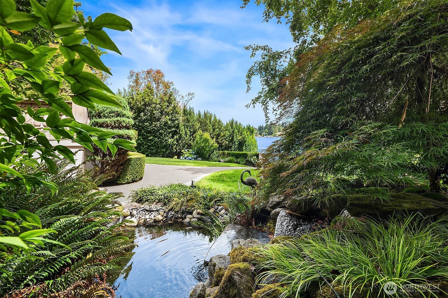 This serene garden scene features a tranquil koi pond with a small waterfall, surrounded by lush ferns, ornamental grasses, and a weeping Japanese maple. A paved pathway winds through the manicured lawn toward a scenic waterfront view in the distance, framed by mature trees and hedges. The perspective is low and immersive, capturing the peaceful atmosphere of a private, well-landscaped estate.