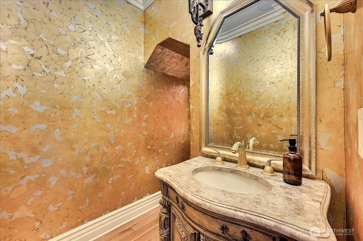 This elegant powder room features a sophisticated vanity with a stone countertop and an ornate, gold-framed mirror. The walls are adorned with a textured, metallic gold and silver faux finish, creating a luxurious and opulent atmosphere. The space is completed by classic gold-toned fixtures and a warm, inviting lighting scheme.