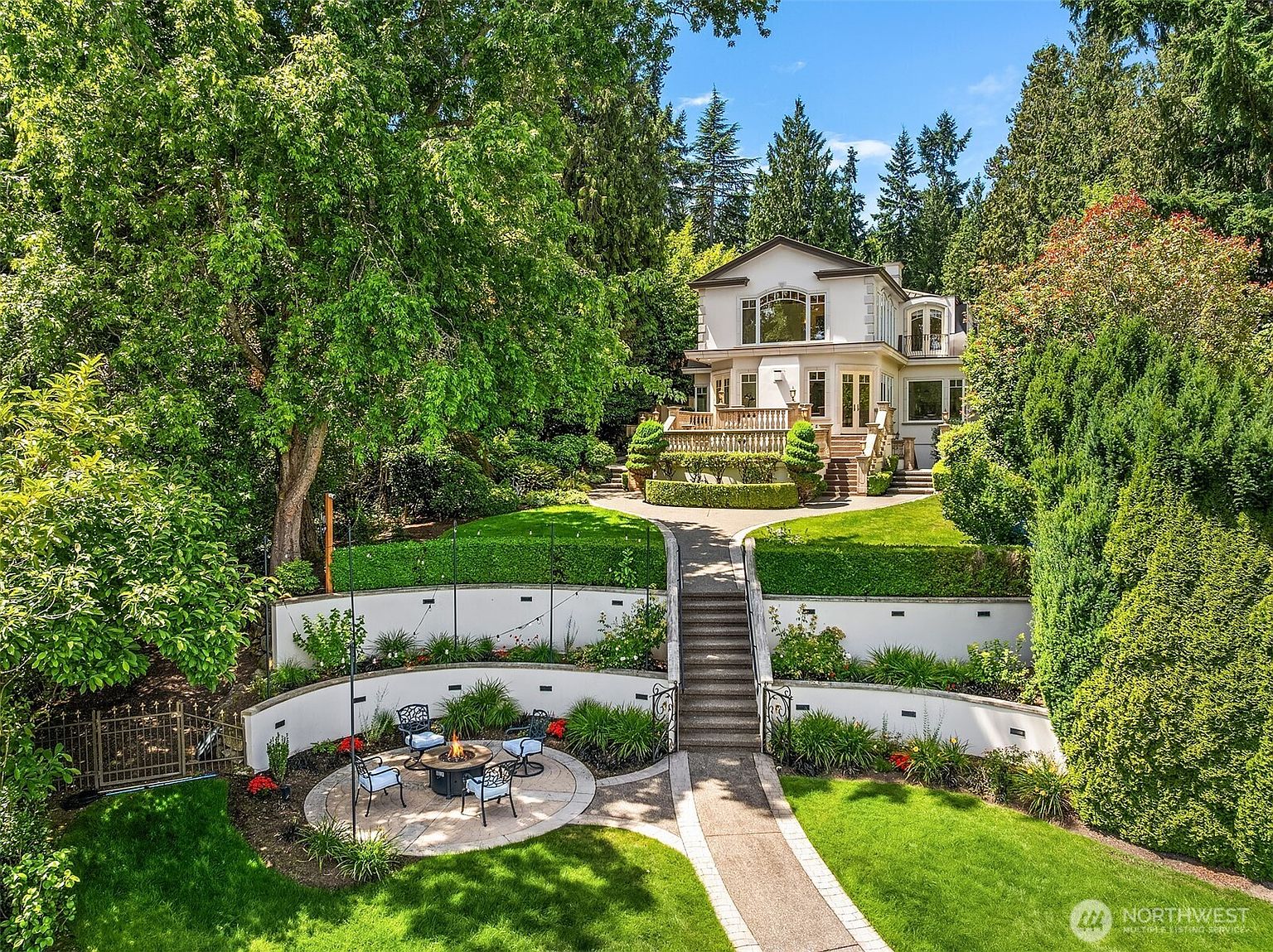 This elevated rear view captures a sophisticated multi-level backyard featuring a stone fire pit area at the base of a terraced retaining wall. A central stone staircase leads up to a manicured lawn and a spacious elevated deck attached to the elegant, light-colored residence. The property is framed by lush, mature trees and greenery, creating a private and serene estate-like atmosphere.