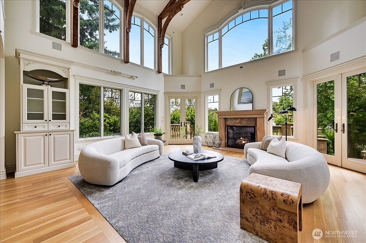 This expansive, light-filled living room features soaring vaulted ceilings with exposed wooden beams and large, floor-to-ceiling windows that offer a panoramic view of the outdoors. The space is anchored by a grand stone fireplace and furnished with two elegant, curved cream-colored sofas arranged around a circular coffee table on a textured area rug. The room exudes a sophisticated, airy atmosphere, blending traditional architectural elements with modern, minimalist furniture.