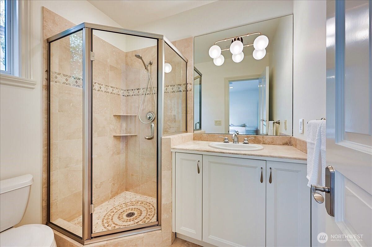 This bright and functional bathroom features a corner glass-enclosed shower with neutral tile work and a built-in shelf. A white vanity with a stone countertop and a large, wall-to-wall mirror creates an open and airy feel. The space is well-lit by a modern multi-bulb light fixture, presenting a clean and well-maintained aesthetic.