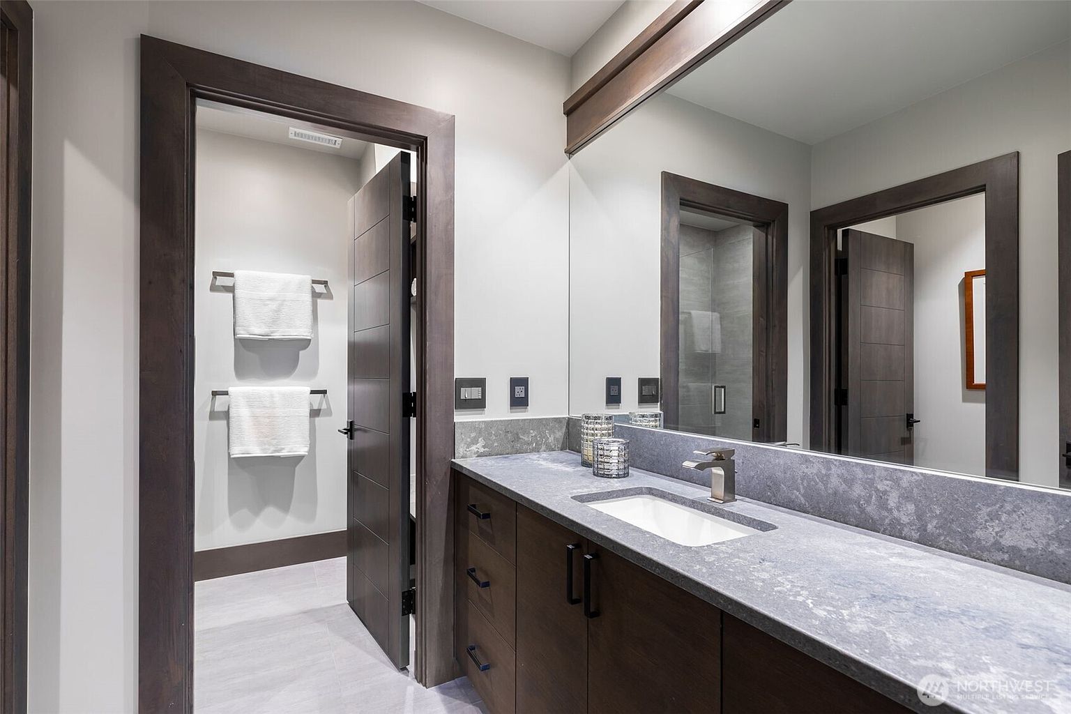 This modern bathroom features a dark wood vanity topped with a grey stone counter and an integrated sink. A large, wide mirror spans the wall above the vanity, reflecting the adjacent doorway and a glimpse of a tiled shower area. The space is finished with neutral walls, dark wood trim, and two white towels hanging on the wall in the doorway area.