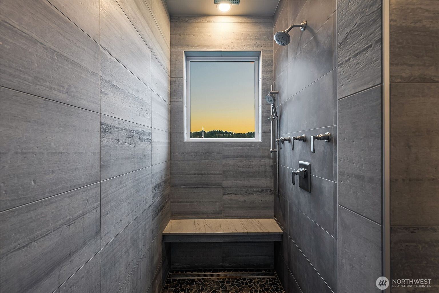 This modern walk-in shower features large-format gray stone tiles on the walls and a textured pebble-style floor. A built-in stone bench sits beneath a window that provides natural light and a scenic view, while a multi-head shower system with a handheld wand is mounted on the side wall. The space is illuminated by a recessed ceiling light, creating a clean and spa-like atmosphere.