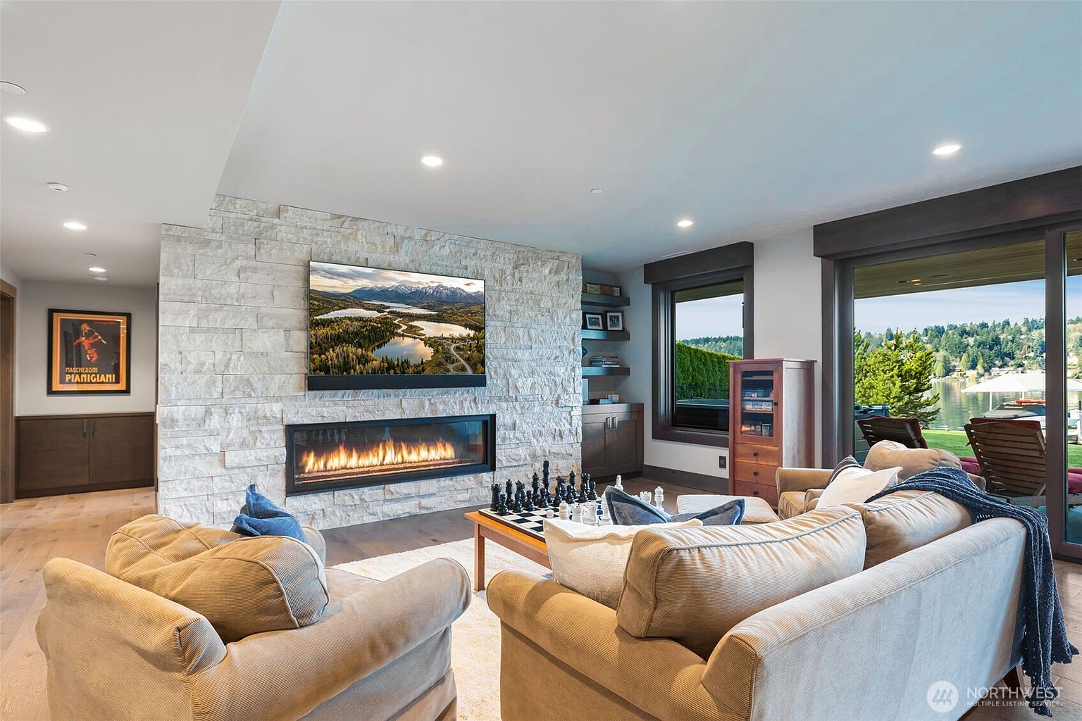 This inviting living room features a prominent stacked-stone chimney breast housing a wide, linear gas fireplace and a mounted television. The space is furnished with comfortable, neutral-toned sofas arranged around a coffee table set up for a game of chess, creating a cozy atmosphere. Large sliding glass doors provide a seamless connection to the outdoors, offering a scenic view of the water and surrounding greenery.