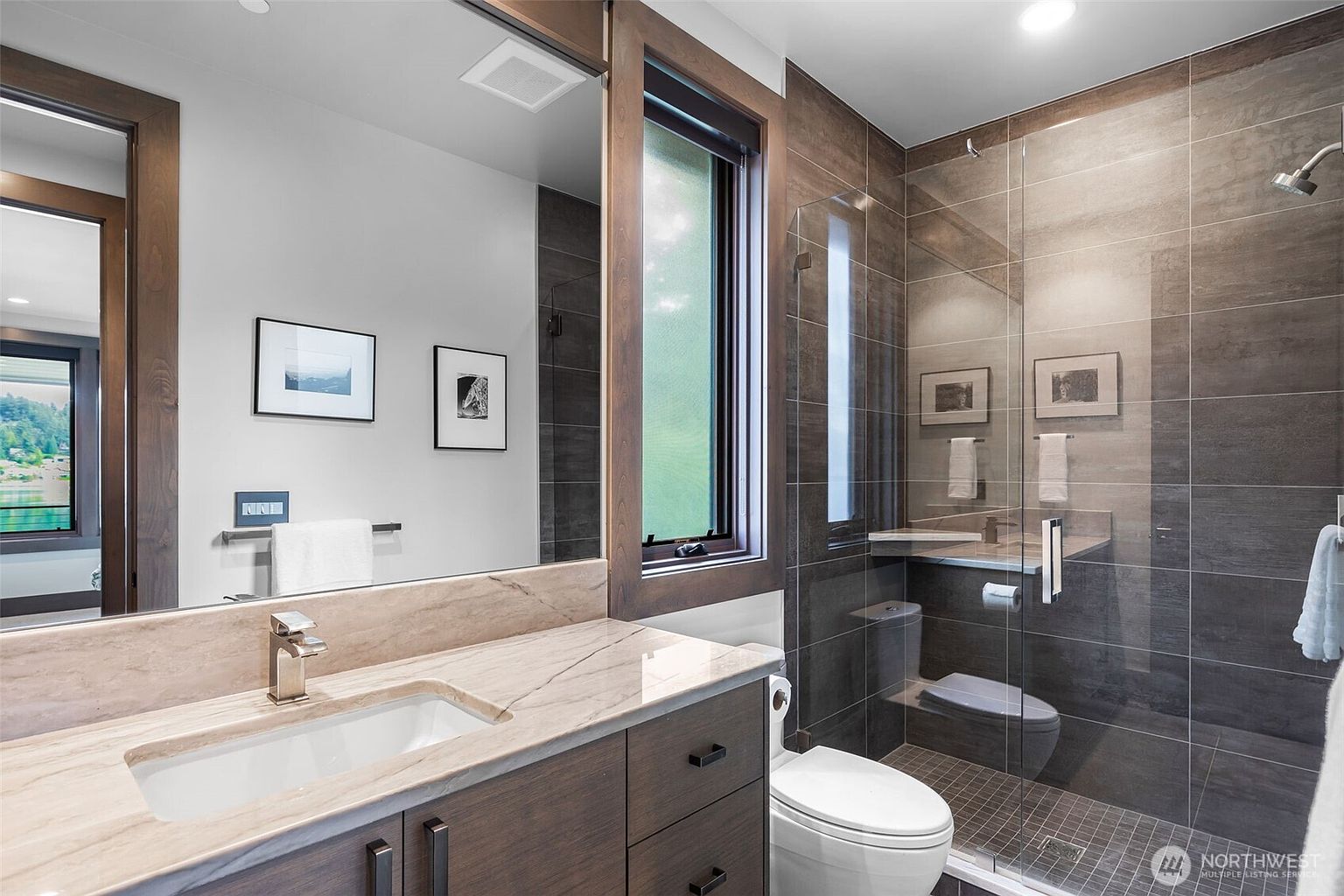 This modern bathroom features a vanity with a stone countertop and a large mirror reflecting the space. To the right, a glass-enclosed shower area includes a toilet and dark, textured wall tiles that create a sophisticated, spa-like atmosphere. The room is well-lit with recessed lighting and includes a tall window that brings in natural light.