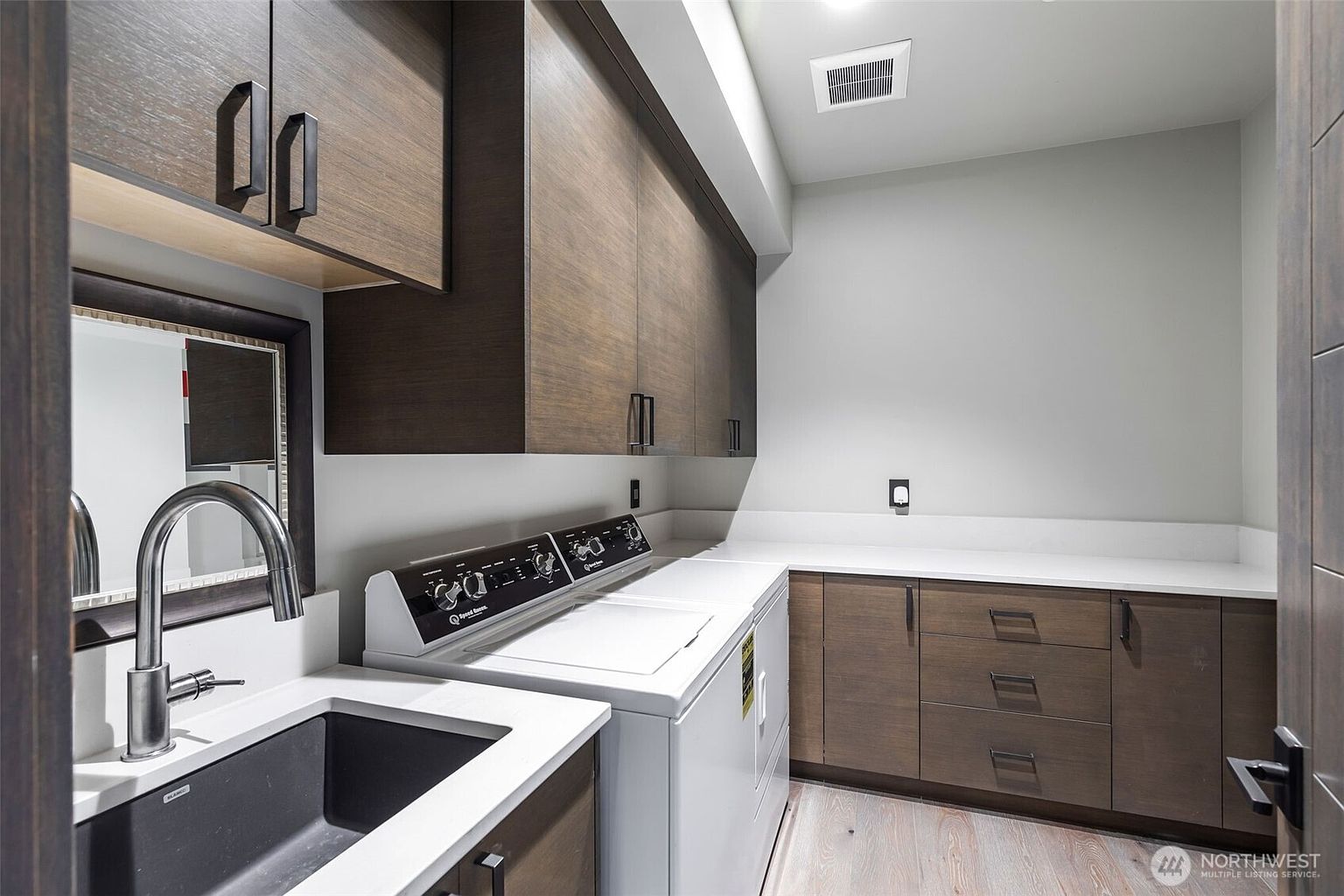 This modern laundry room features dark wood cabinetry, a white countertop, and a built-in sink with a sleek faucet. A white washer and dryer set are positioned centrally, complemented by a light-colored wood-look floor. The space is well-lit with recessed lighting and offers a clean, functional design with ample storage.