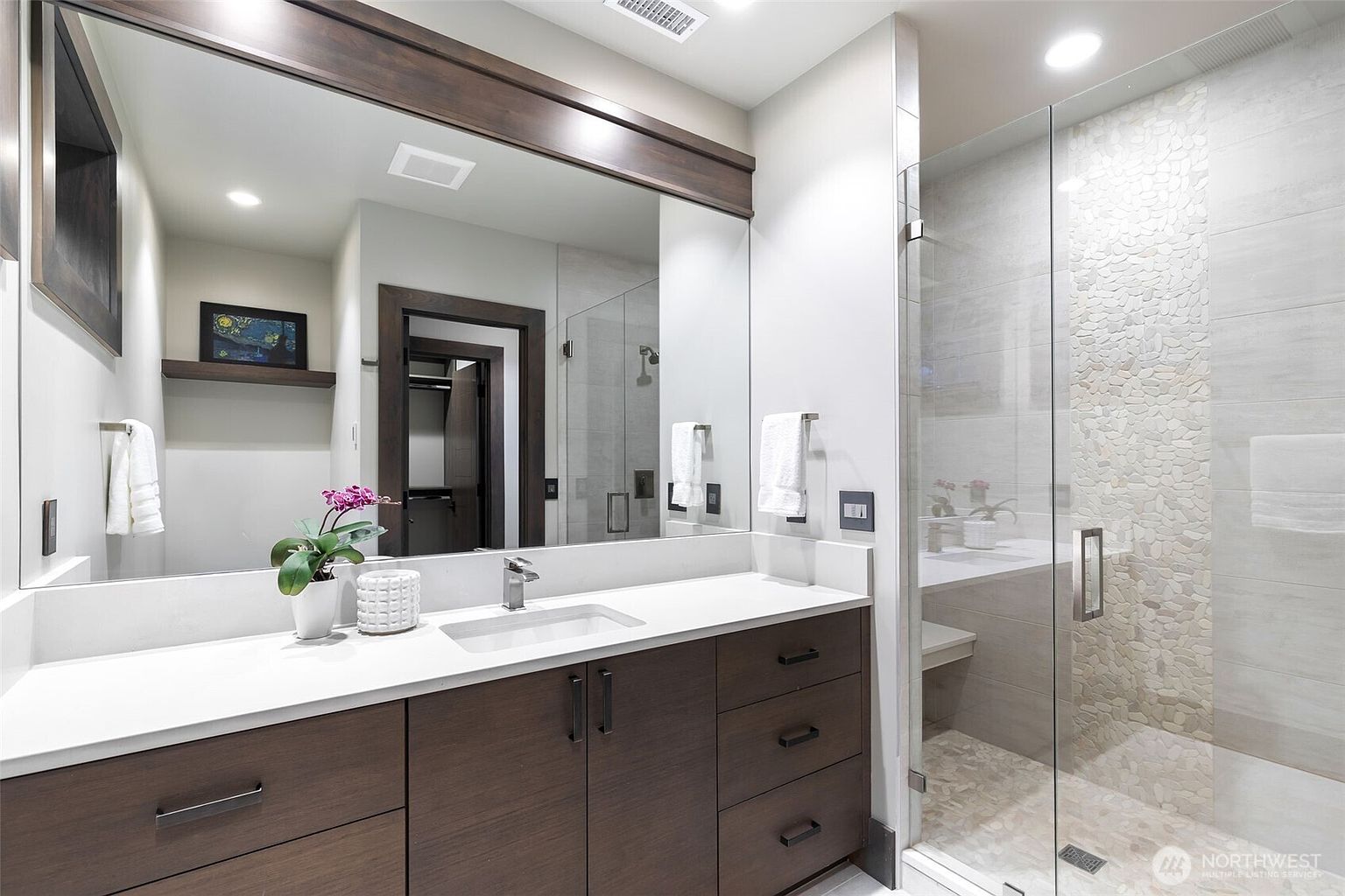 This modern bathroom features a dark wood vanity with a white countertop and a large, wide mirror that reflects the adjacent walk-in closet. To the right, a glass-enclosed shower showcases a textured stone accent wall and a built-in bench. The space is bright and clean, utilizing a neutral color palette and contemporary fixtures to create a spa-like atmosphere.