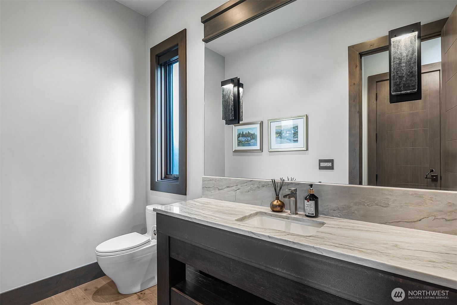 This modern bathroom features a dark wood vanity topped with a light-toned stone countertop and an integrated sink. A large, frameless mirror spans the wall above the vanity, flanked by two contemporary wall-mounted light fixtures, while two small framed artworks add a decorative touch. To the left, a white toilet is positioned near a tall, narrow window that allows natural light to enter the space.