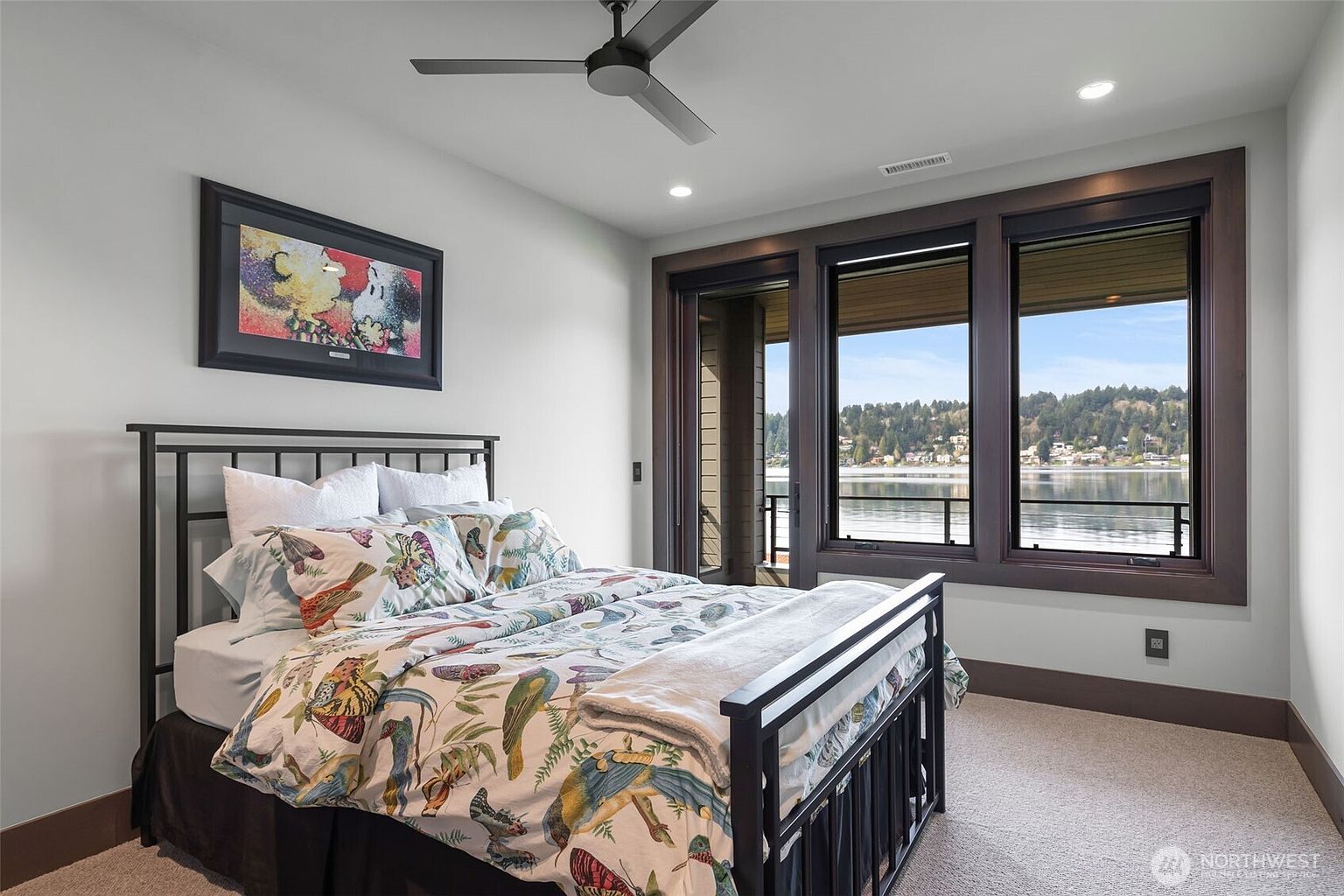 This bedroom features a dark metal bed frame dressed in a vibrant, nature-inspired patterned duvet and a neutral throw blanket. A large piece of colorful artwork hangs above the headboard, while a modern ceiling fan provides overhead lighting. The room is anchored by a set of large windows that offer a clear, expansive view of the lake and surrounding landscape.