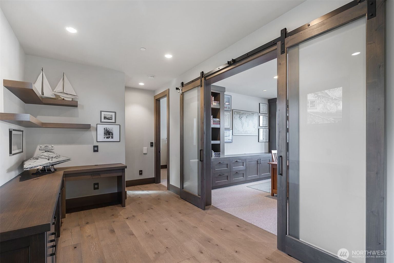 This sophisticated home office area features light wood flooring and a custom built-in desk workspace adorned with model ships and a detailed starship replica. A pair of sliding barn-style doors with frosted glass panels separates this area from an adjacent study, which is furnished with dark cabinetry, a desk, and framed wall art. The space is illuminated by recessed lighting, creating a professional and organized atmosphere.