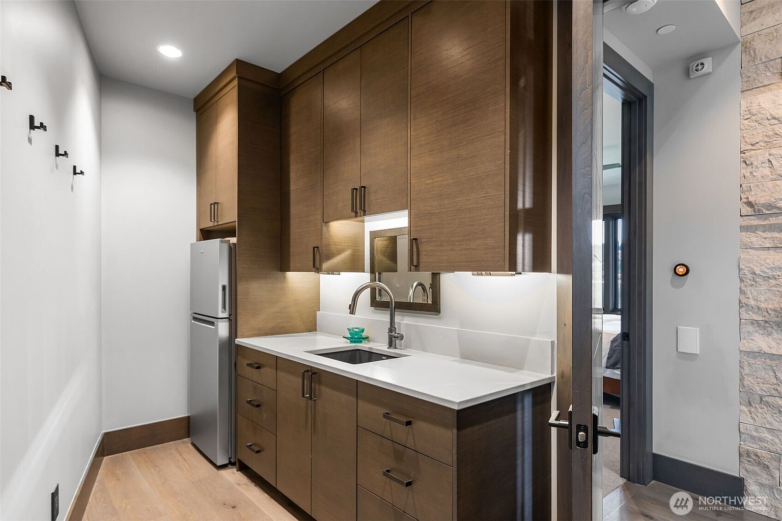 This compact kitchenette features warm-toned wood cabinetry, a sleek white countertop, and an integrated stainless steel refrigerator. A modern faucet sits above a small sink, complemented by under-cabinet lighting that highlights the workspace. The area is adjacent to a doorway leading into a bedroom, with a stone-clad wall visible on the right, creating a sophisticated and functional transition space.
