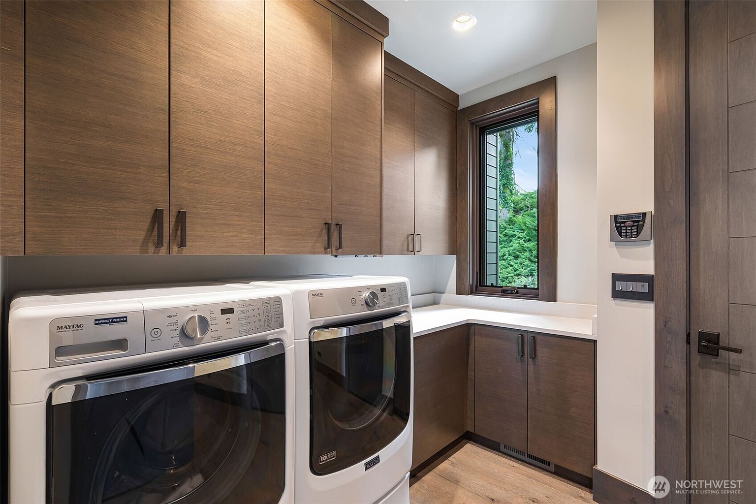 This laundry room features a clean, modern aesthetic with wood-toned cabinetry and white appliances. A white countertop provides workspace above the lower cabinets, while a window allows natural light to brighten the area. The space is efficiently designed with a neutral color palette and contemporary hardware.