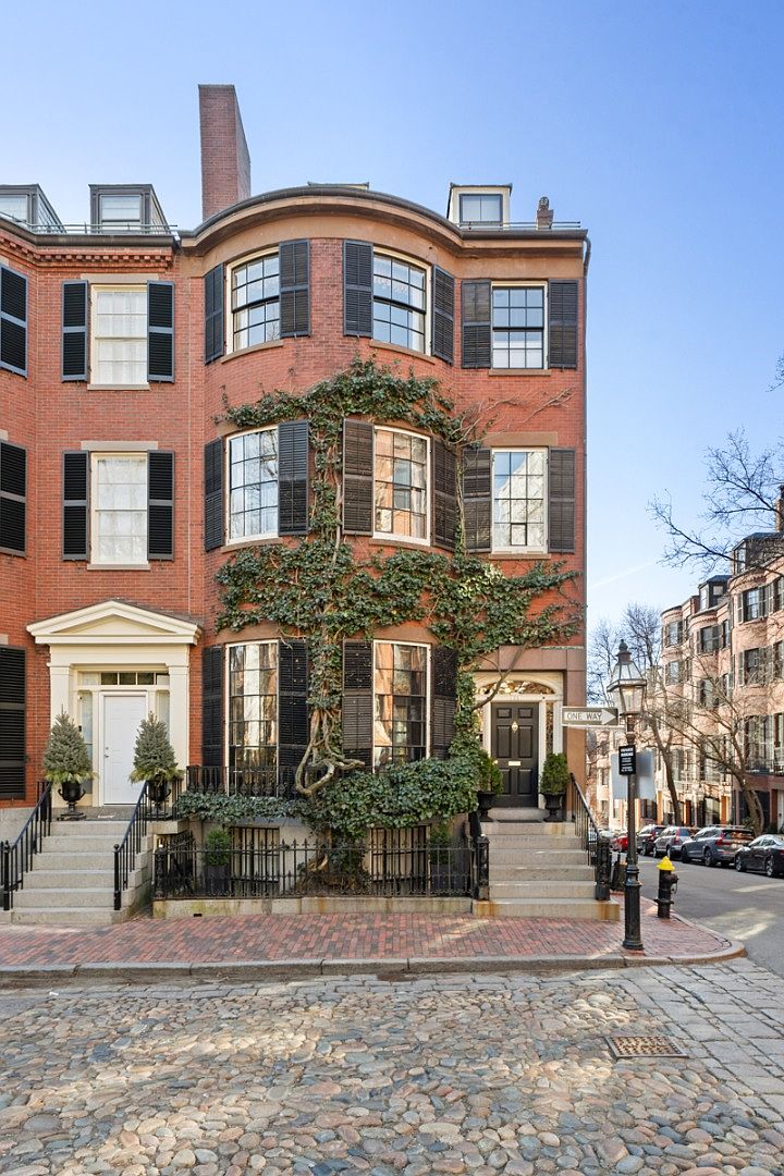 This is a front view of a classic brick townhouse with black shutters and a curved facade. The building features two separate entryways with stoops and wrought iron railings. Ivy climbs the front of the building, adding a touch of greenery and character to the property.