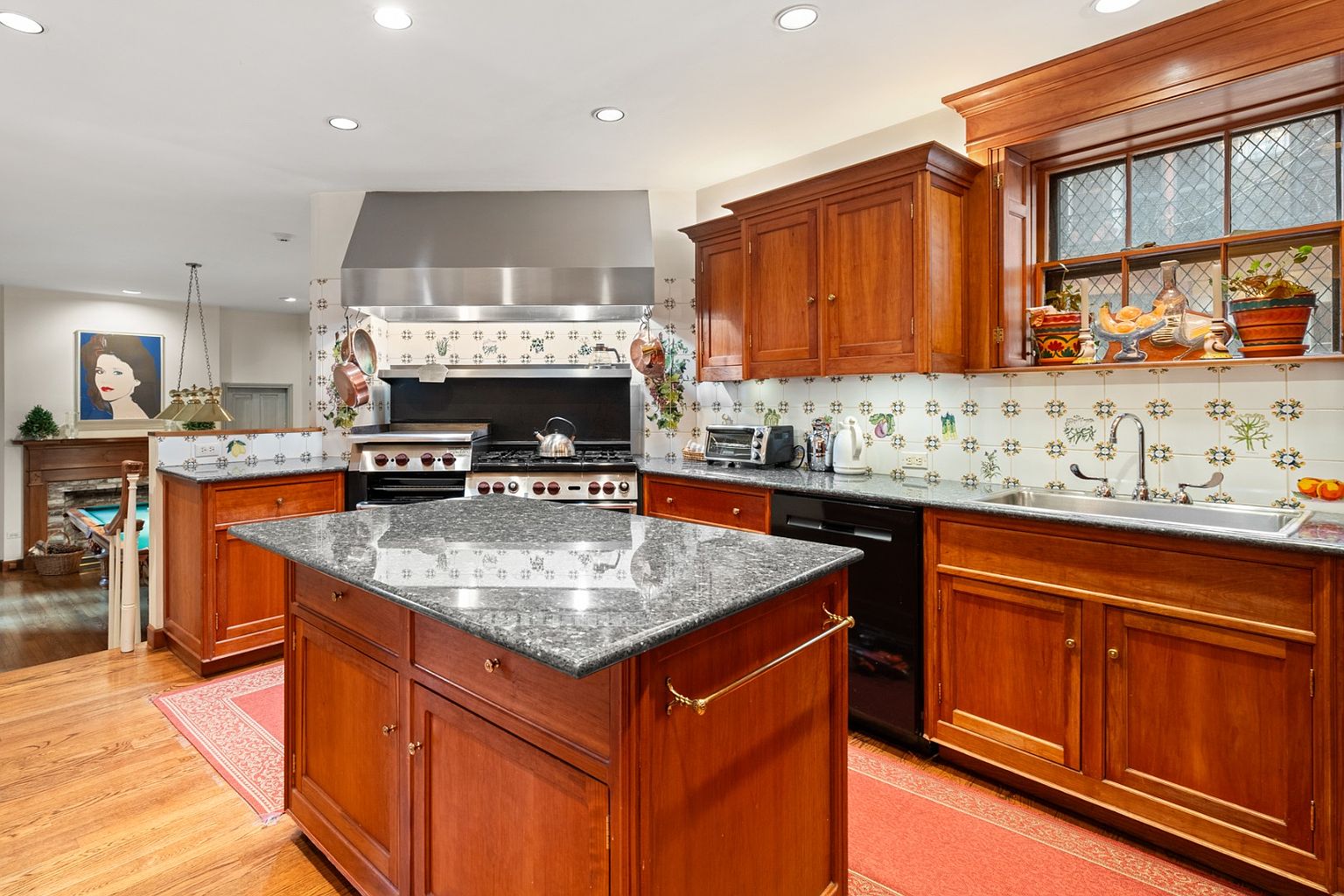 This is a warm and inviting kitchen featuring custom wood cabinetry, granite countertops, and a stainless steel range hood. The kitchen island provides ample workspace, and the decorative tile backsplash adds a touch of charm. The room is well-lit with recessed lighting, and the hardwood floors contribute to the overall cozy atmosphere.