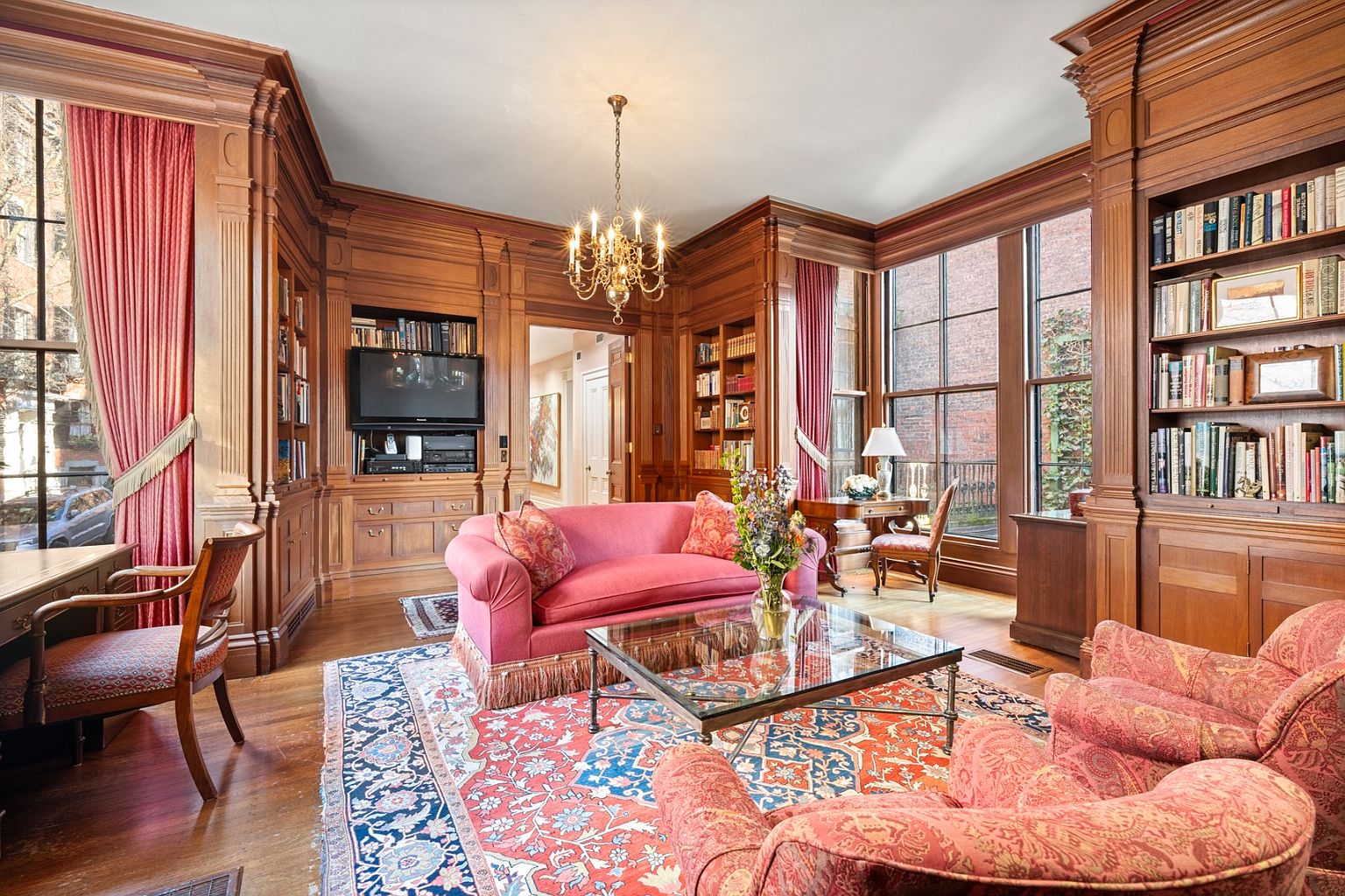 This is a luxurious living room featuring rich wood paneling, built-in bookshelves, and a classic chandelier. A vibrant pink sofa and patterned armchairs add a pop of color to the room, which is grounded by an ornate area rug. Large windows provide ample natural light, creating a warm and inviting atmosphere.