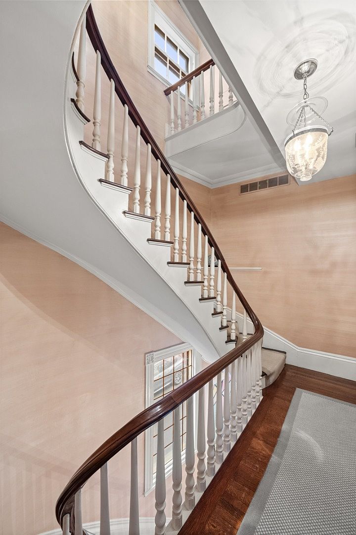 This interior shot showcases a curved staircase with white balusters and a dark wood handrail. The walls are covered in a textured, light peach wallpaper, and a decorative light fixture hangs from the ceiling. A window provides natural light, and a glimpse of the hardwood floor and a rug can be seen at the bottom of the stairs, creating an elegant and inviting atmosphere.