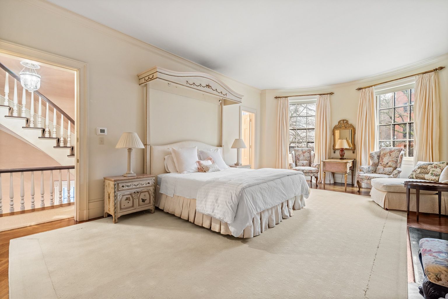 This is a spacious primary bedroom featuring a four-poster bed with white linens, flanked by matching nightstands and lamps. The room has a neutral color palette, large windows with elegant draperies, and a sitting area with upholstered chairs. The overall impression is one of luxury and comfort.