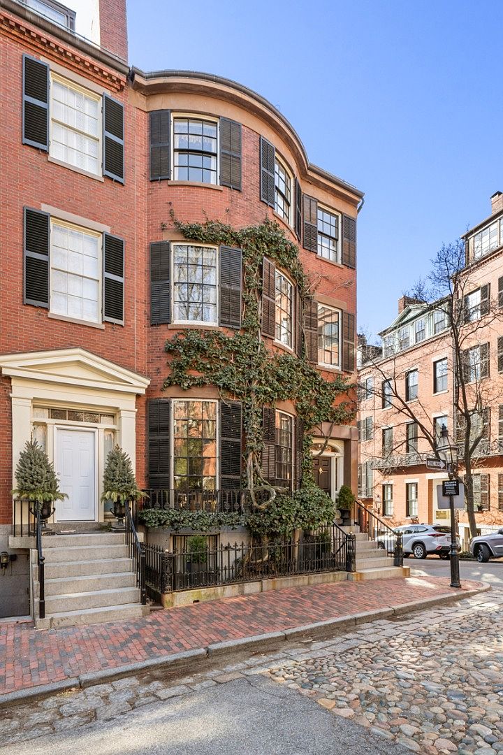 This is a front view of a classic brick townhouse with black shutters and a curved facade. The entrance features a white door with a decorative pediment, flanked by small evergreen trees. Ivy climbs the side of the building, adding a touch of nature to the urban setting. The street is paved with cobblestones, enhancing the historic charm.