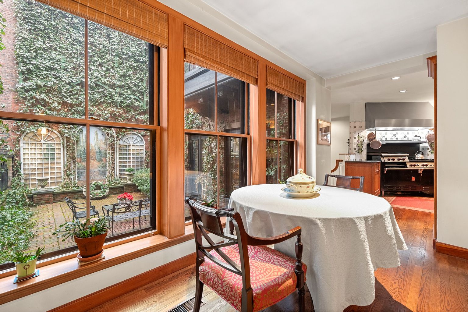 This image showcases a charming dining area with a round table draped in a white tablecloth, complemented by an elegant chair with patterned upholstery. Large windows offer a view of a lush courtyard, enhancing the room's appeal. The kitchen is visible in the background, featuring a classic black range and wooden cabinetry, adding to the home's character.