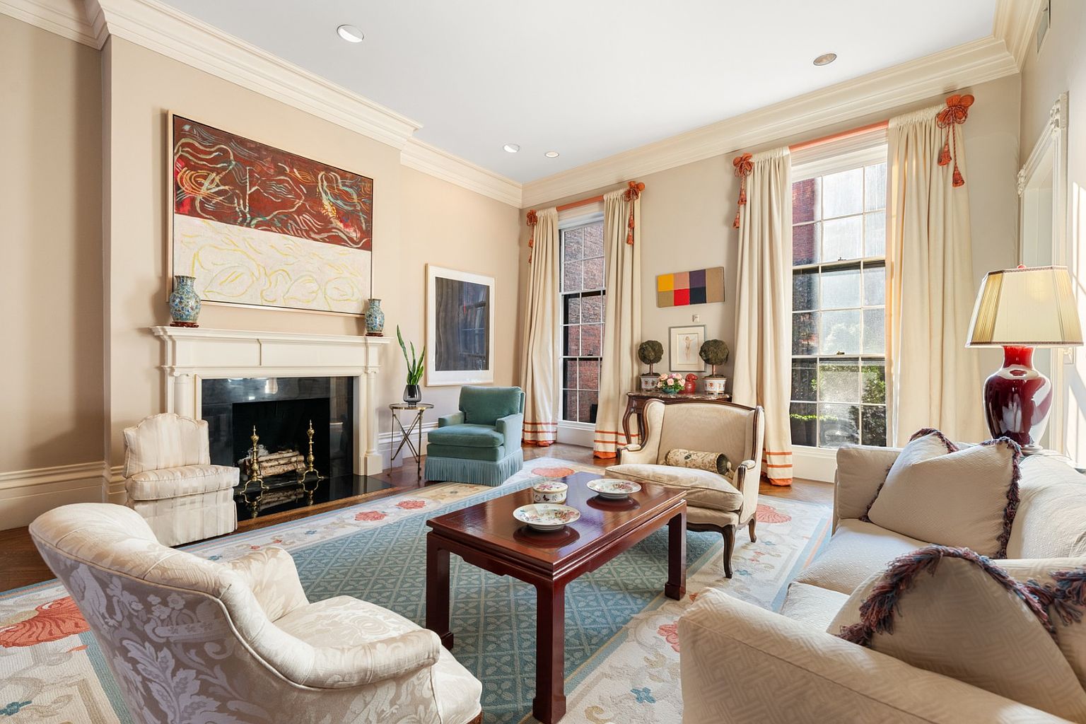 This is a well-appointed living room featuring a fireplace with an ornate mantel, a large abstract painting above it, and several comfortable armchairs and a sofa arranged around a dark wood coffee table. The room is decorated in neutral tones with pops of color in the artwork and accessories, creating a sophisticated and inviting atmosphere. Large windows provide ample natural light, enhancing the room's warmth and elegance.