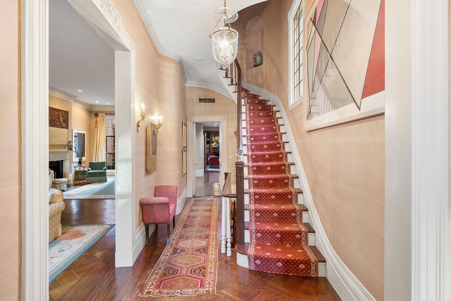 This interior shot showcases a grand hallway with a staircase. The hallway features hardwood floors in a herringbone pattern, a patterned runner rug, and light peach-colored walls. A staircase with a red patterned carpet leads to the upper level, illuminated by a chandelier. Artwork adorns the walls, and doorways lead to other rooms, creating a sense of depth and elegance.