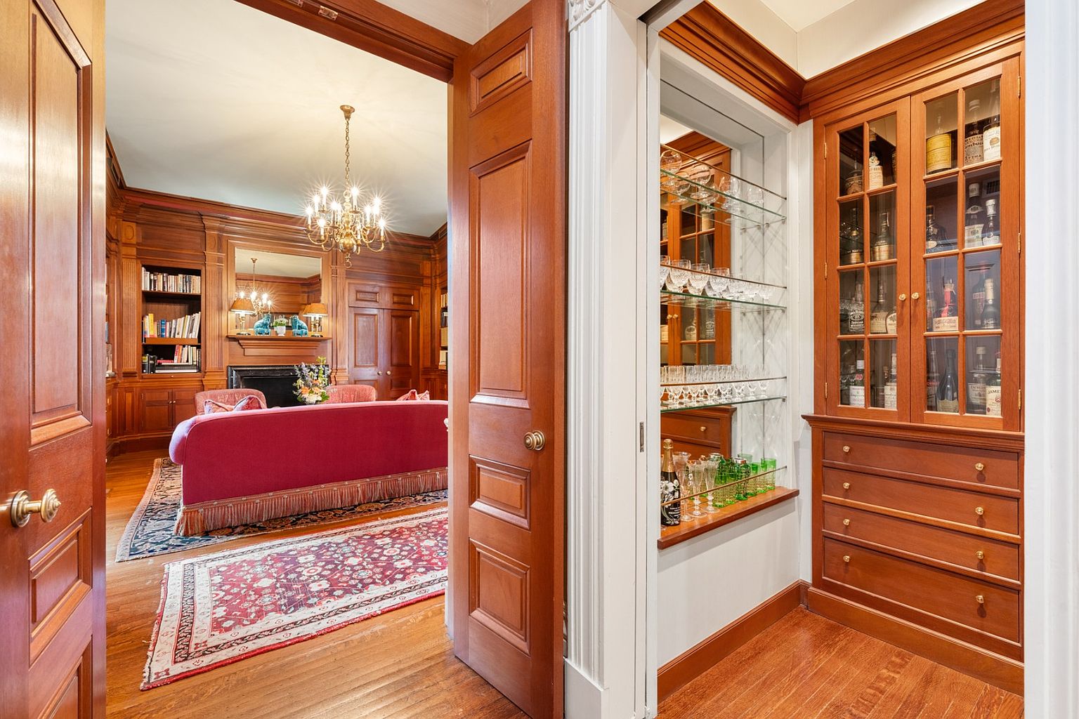This image showcases a luxurious living room viewed through an open doorway, featuring rich wood paneling, a chandelier, and a red sofa with fringe detailing. Adjacent to the living room is a built-in bar area with glass shelves and a cabinet displaying various bottles, suggesting a sophisticated and elegant home environment. The hardwood floors and patterned rug add to the room's classic charm.