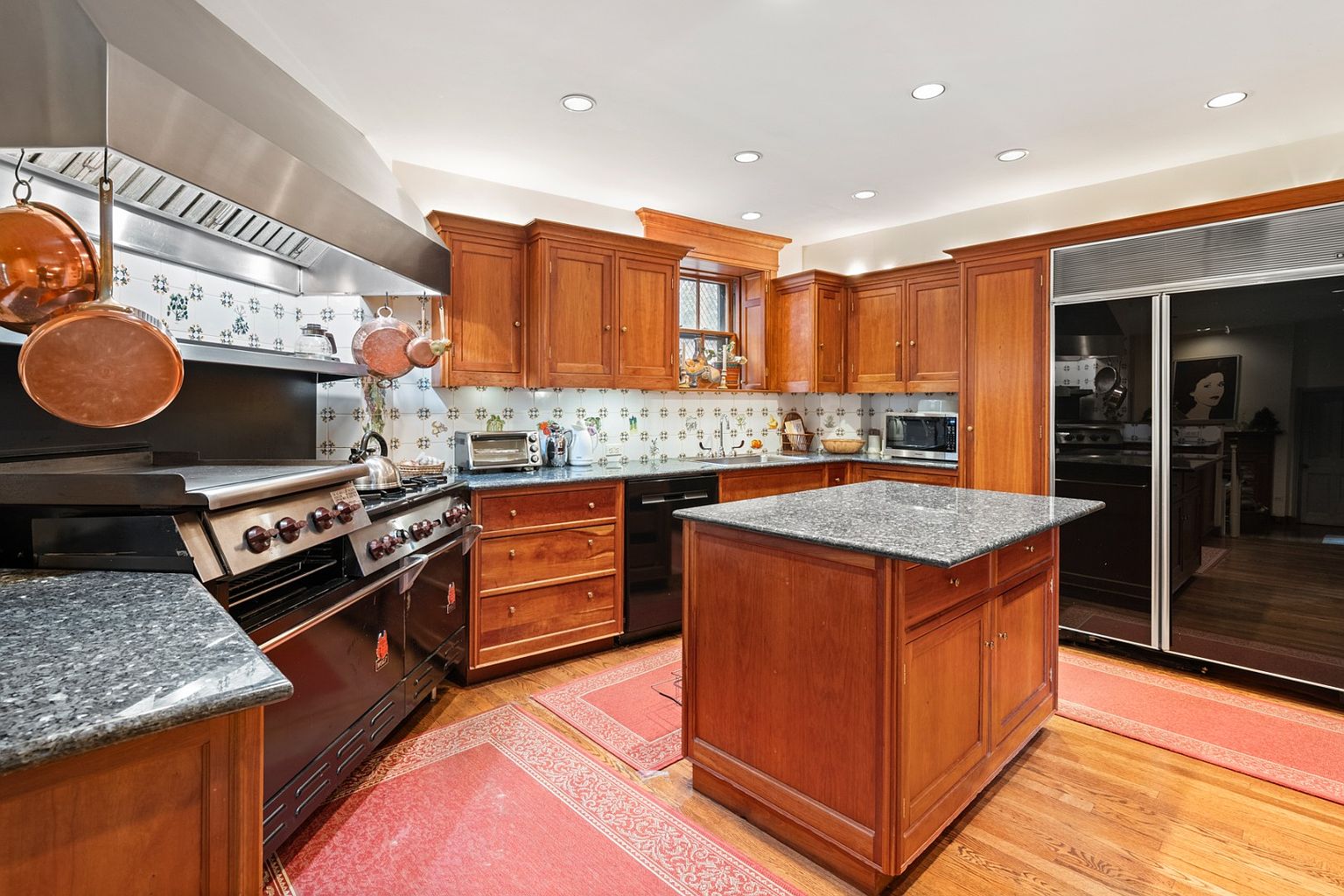 This is a warm and inviting kitchen featuring wooden cabinetry, granite countertops, and a large professional-grade range with a stainless steel hood. A central island provides additional workspace, and the patterned backsplash adds a touch of character. The overall impression is one of a well-equipped and comfortable space for cooking and entertaining.