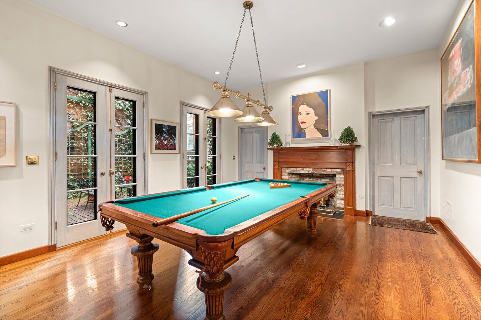 This interior shot showcases a well-appointed living room featuring a classic billiard table as the focal point. The room is illuminated by a vintage-style chandelier, and the hardwood floors add warmth. French doors lead to an outdoor space, and artwork adorns the walls, contributing to an elegant and inviting atmosphere.