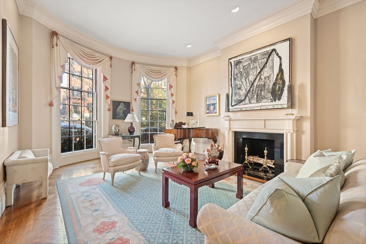 This is an elegant living room featuring a fireplace with a large abstract painting above it, two armchairs, and a sofa with light blue pillows. The room has a bay window with decorative curtains, a piano, and a patterned area rug. The overall impression is one of sophistication and comfort.