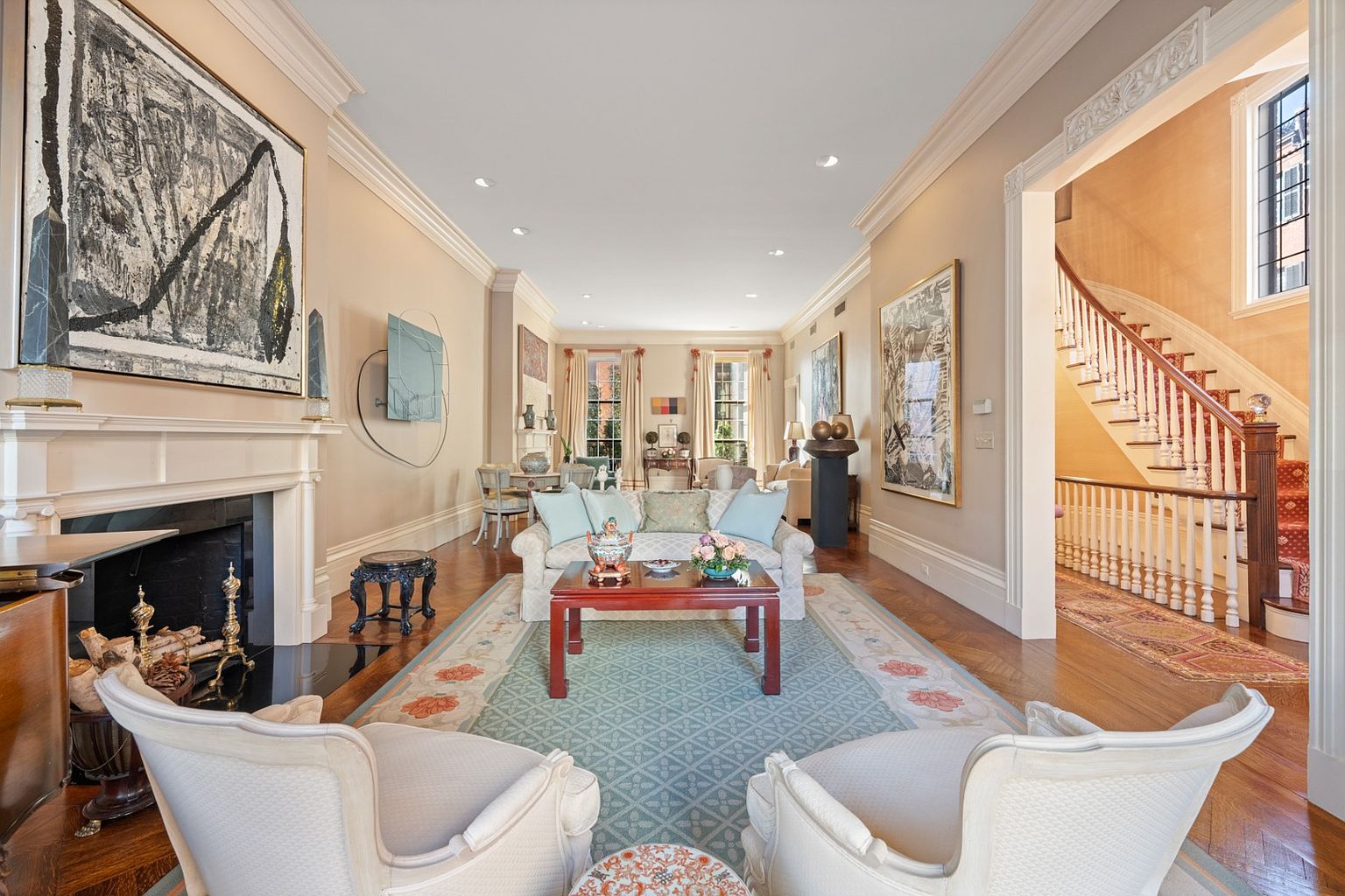 This is an interior shot of a luxurious living room featuring elegant furnishings, including a plush sofa, armchairs, and a decorative coffee table. The room is adorned with artwork and a fireplace, creating a sophisticated and inviting atmosphere. A glimpse of the staircase adds to the grandeur of the home.