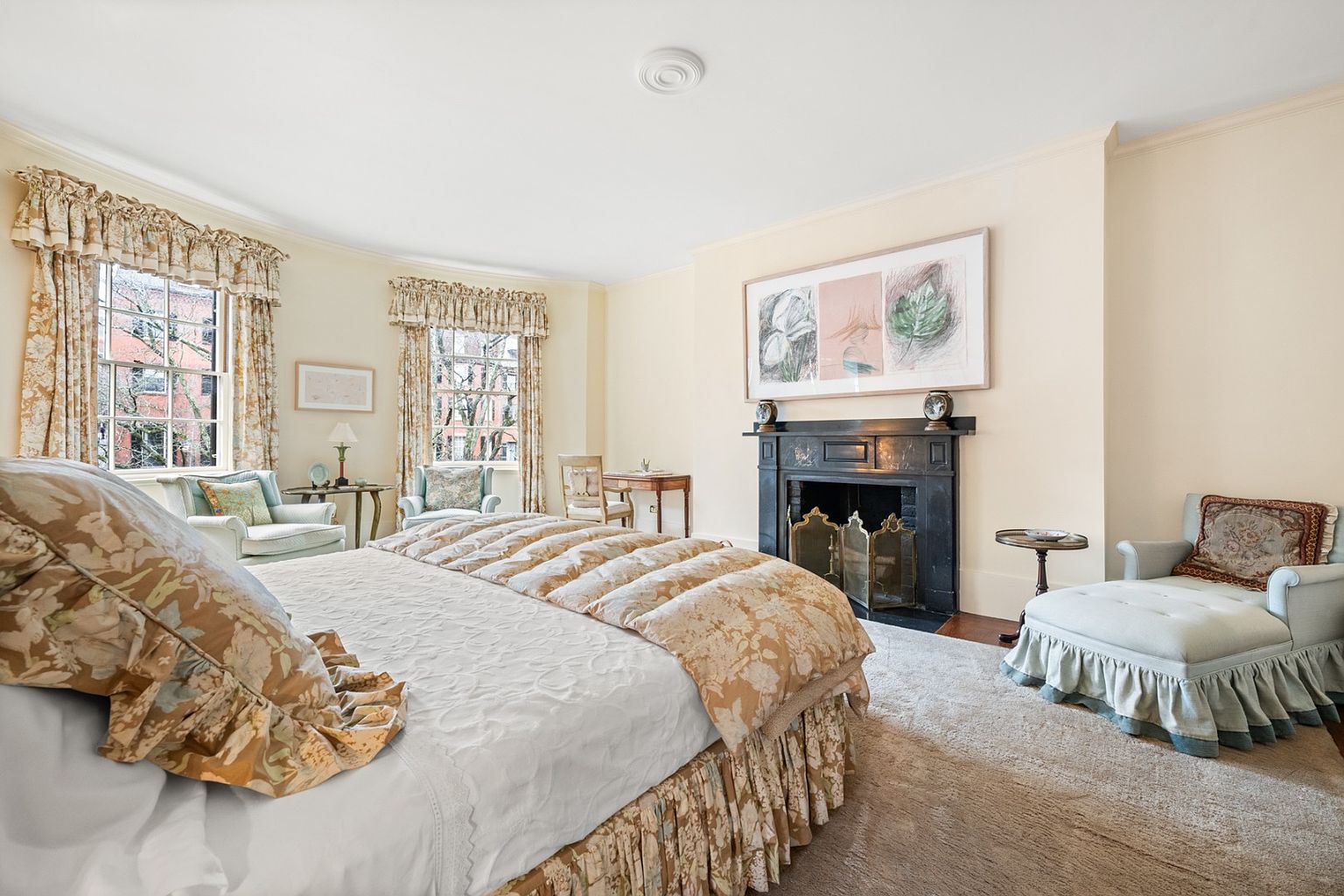 This is a well-lit primary bedroom featuring a large bed with floral patterned bedding, two windows with matching floral curtains, and a black fireplace with an ornate screen. A chaise lounge sits to the right of the fireplace, and the room is decorated in a traditional style with neutral wall colors and a plush carpet, creating a comfortable and inviting atmosphere.