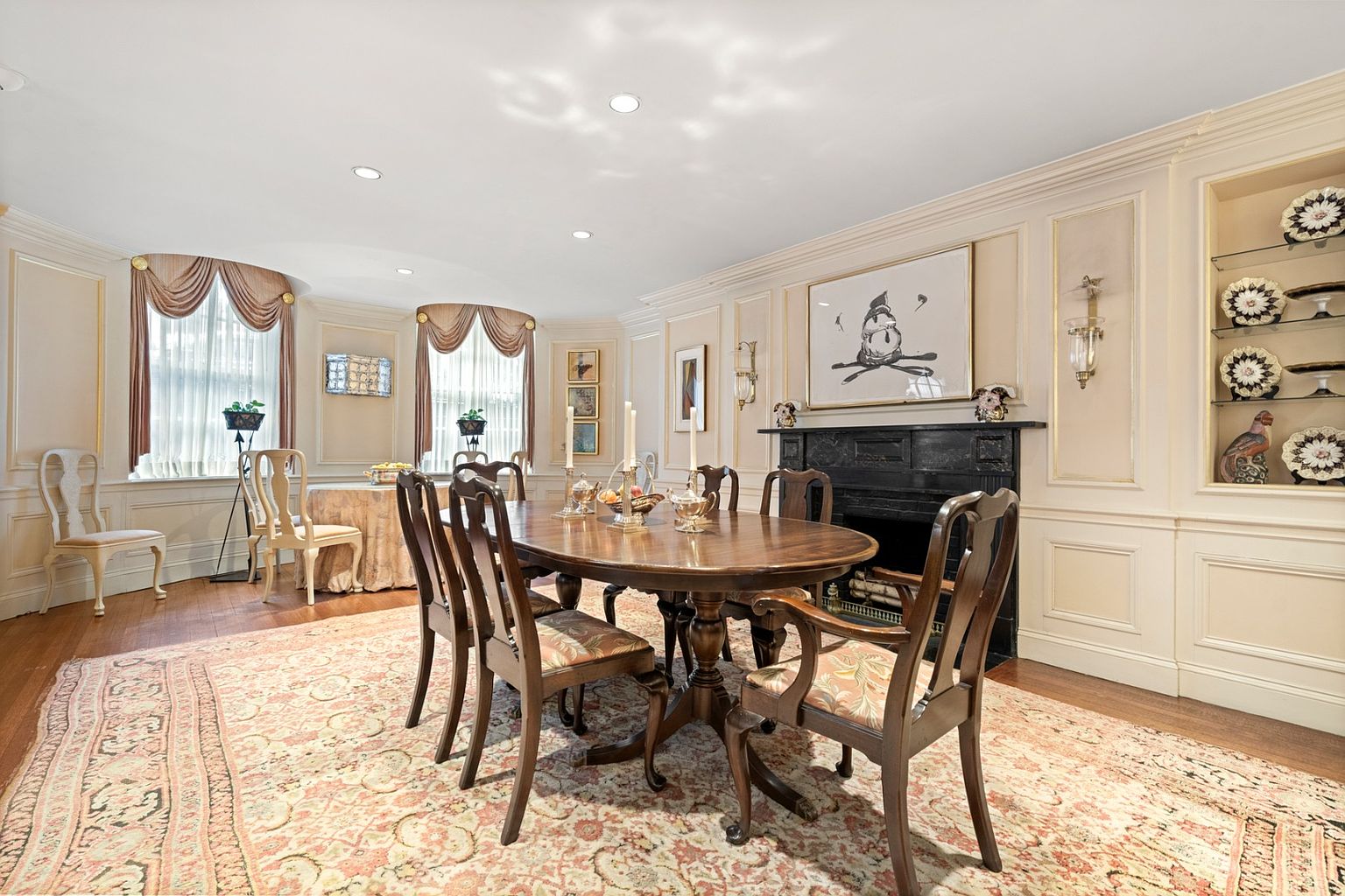 This is an elegant dining room featuring a large oval wooden table surrounded by upholstered chairs, set upon an ornate rug. A black fireplace with a decorative mantle and artwork above it adds a focal point, while built-in shelving displays decorative plates and a ceramic bird. The room is well-lit with natural light from the windows and recessed lighting, creating a warm and inviting atmosphere.