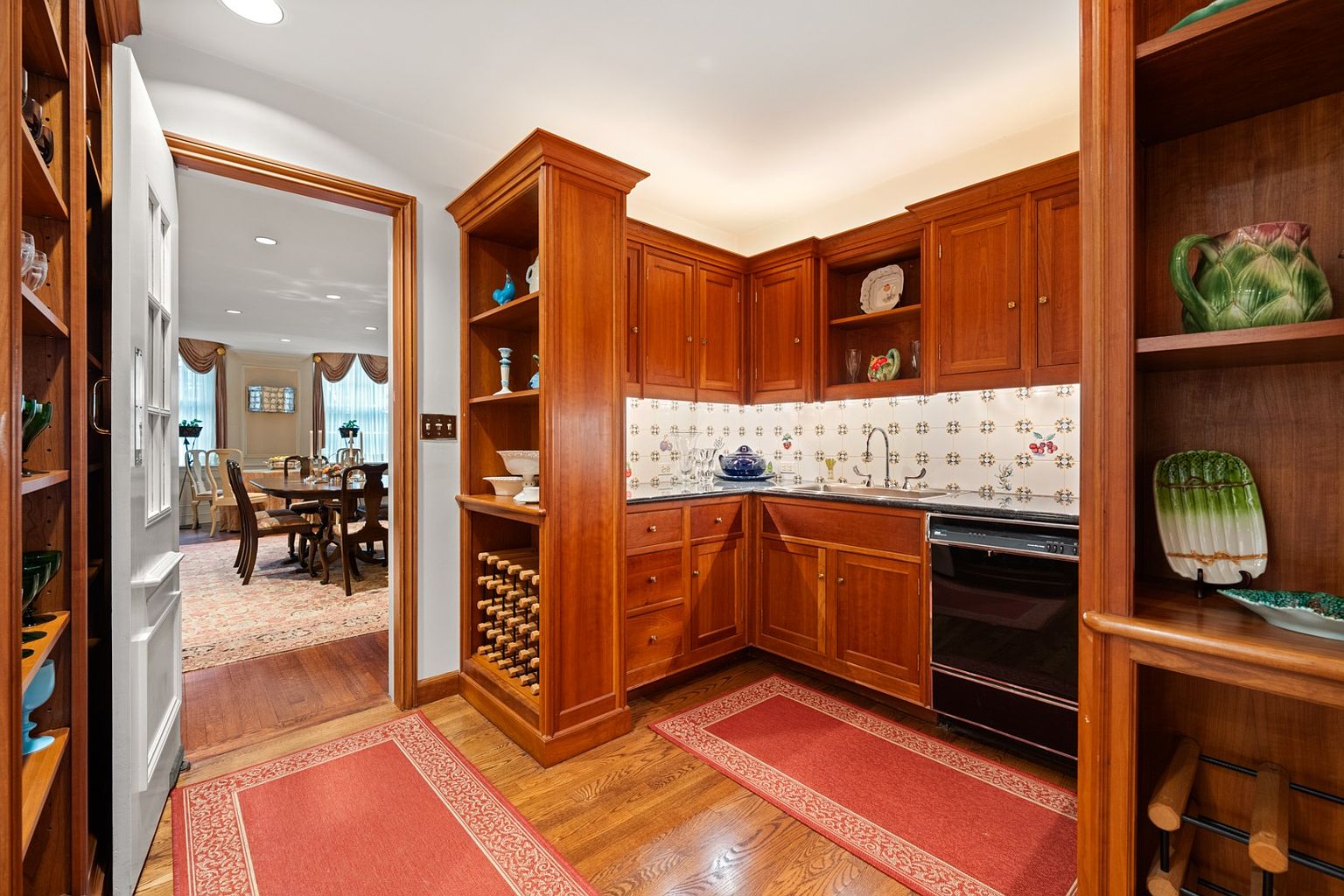 This is a warm and inviting kitchen featuring rich wood cabinetry and shelving, complemented by a patterned tile backsplash. The kitchen is well-lit and includes a black dishwasher, with a glimpse into a dining area through an open doorway. Red accent rugs add a pop of color to the hardwood floors, creating a cozy and classic atmosphere.