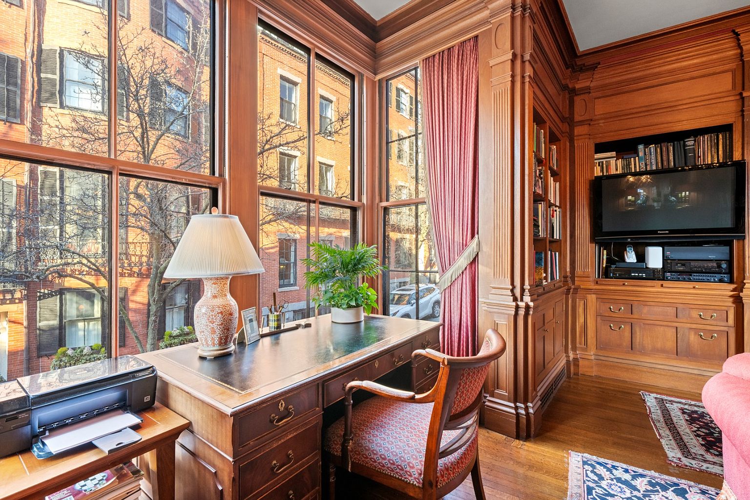 This is an interior shot of a well-appointed office or study. The room features rich wood paneling, built-in bookshelves, and a large window offering a view of a brick building and trees. A wooden desk with a green leather top sits in front of the window, complemented by a patterned chair, creating a sophisticated and inviting workspace.