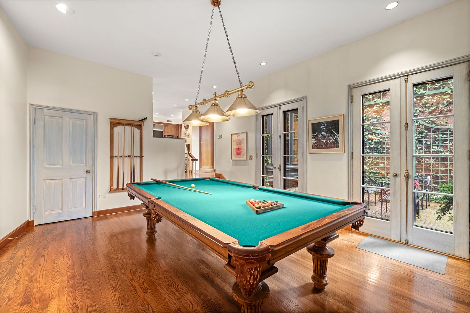 This interior shot showcases a well-appointed living room featuring a classic billiard table as its centerpiece. The room is illuminated by a vintage-style pendant light fixture hanging above the table, and natural light streams in through French doors that lead to an outdoor space. The hardwood floors and neutral wall colors create a warm and inviting atmosphere.
