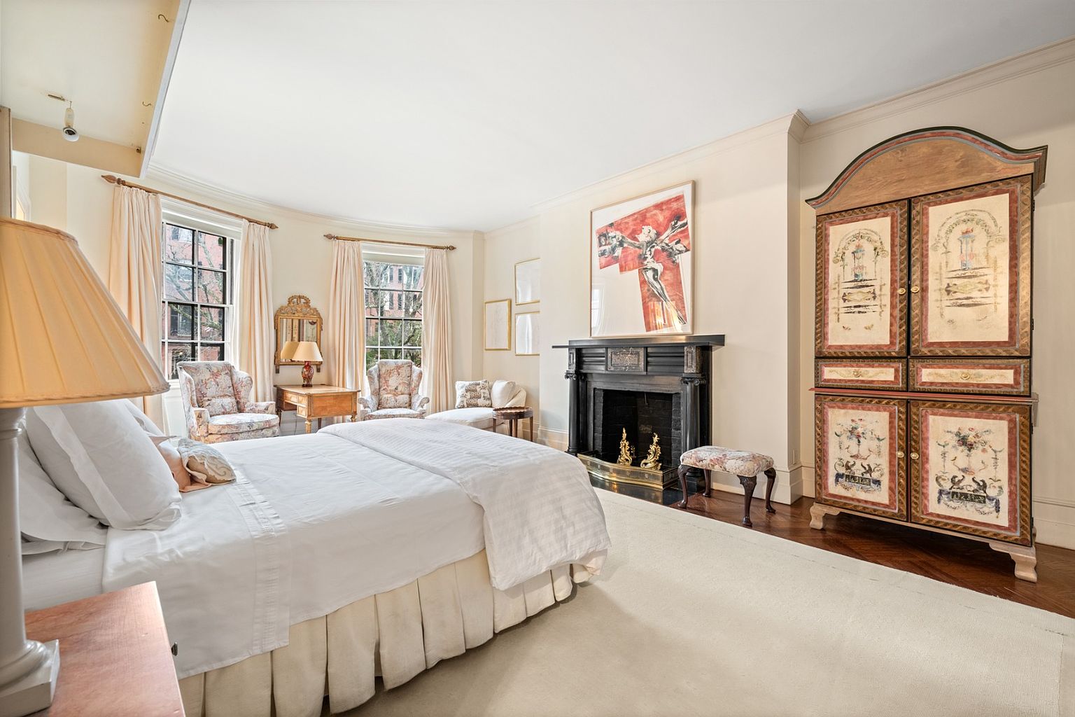 This is a primary bedroom featuring a large bed with white linens, a fireplace with a dark mantel, and an ornate armoire. The room has a traditional style with neutral walls, hardwood floors partially covered by a light-colored rug, and windows with light-colored curtains. The perspective is from the side of the bed, showcasing the room's depth and features.