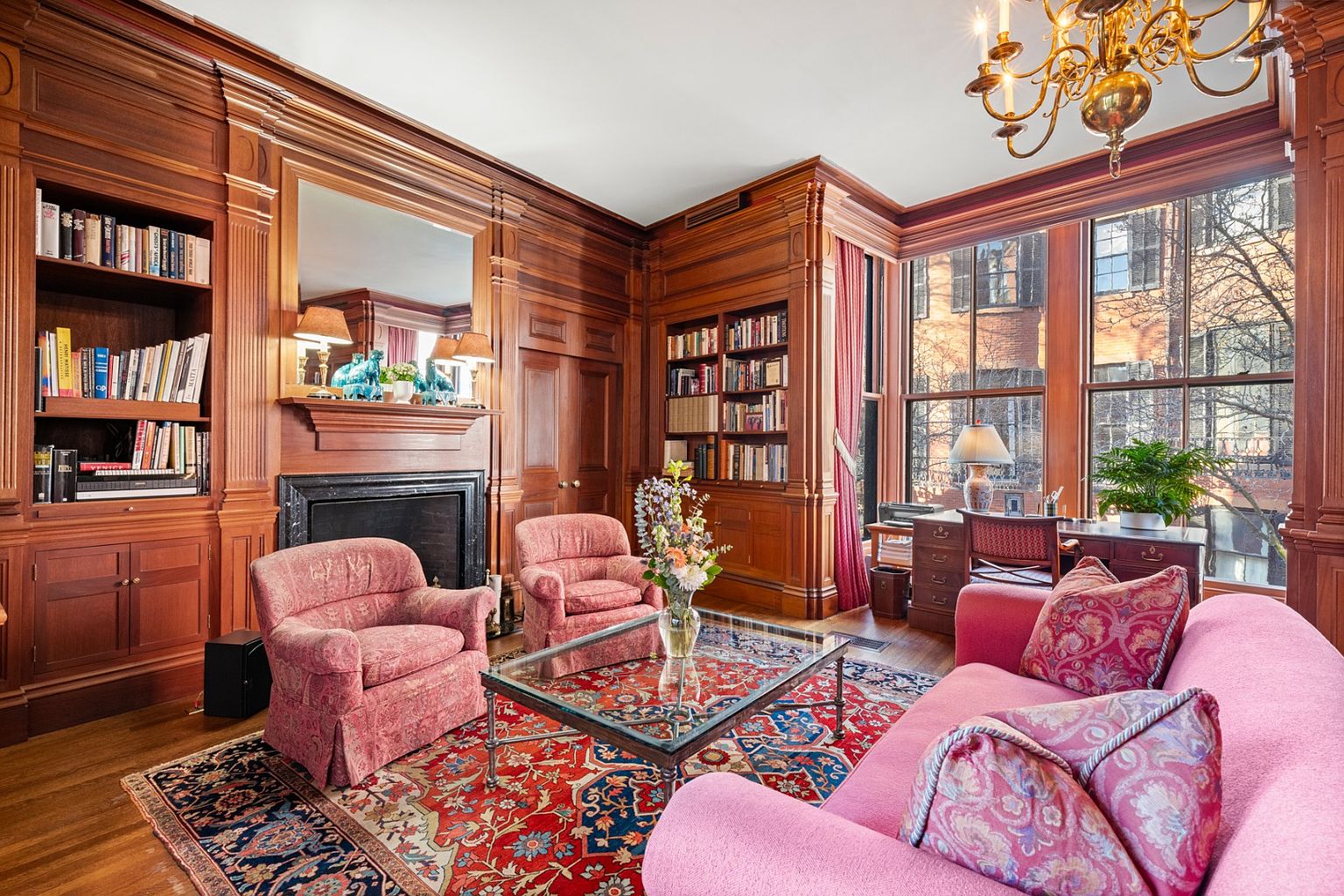 This is a classic living room featuring rich wood paneling, built-in bookshelves, and a fireplace with a decorative mantel. The room is furnished with a pink sofa, patterned armchairs, and a glass-topped coffee table, all set upon an ornate area rug. Large windows provide natural light, and a chandelier hangs from the ceiling, creating a warm and inviting atmosphere.