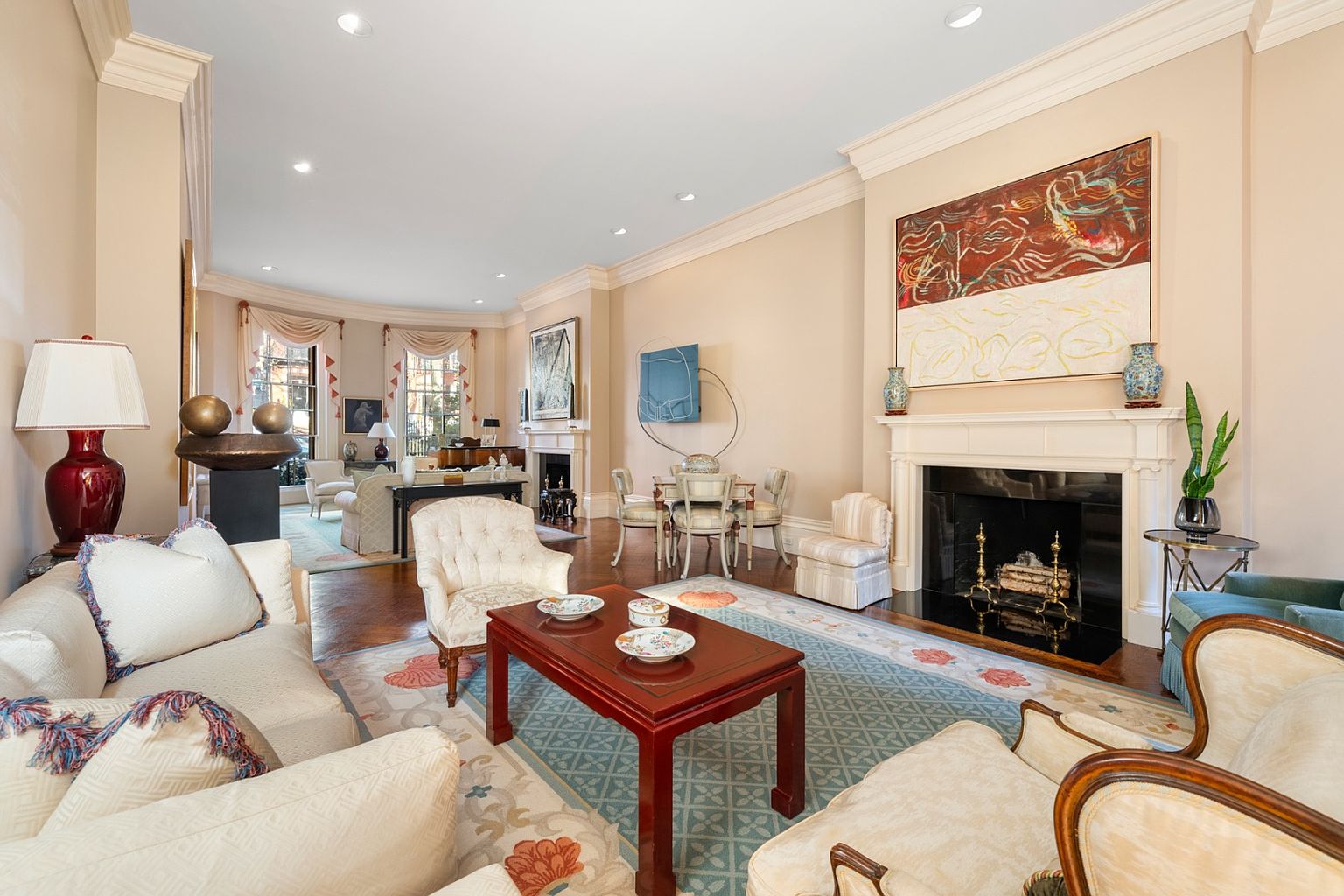 This is a spacious and elegant living room featuring a mix of classic and artistic elements. The room is furnished with plush sofas, armchairs, and a dark wood coffee table, all arranged on a patterned rug. A fireplace with a large painting above it serves as a focal point, complemented by decorative vases and a plant, creating a sophisticated and inviting atmosphere.
