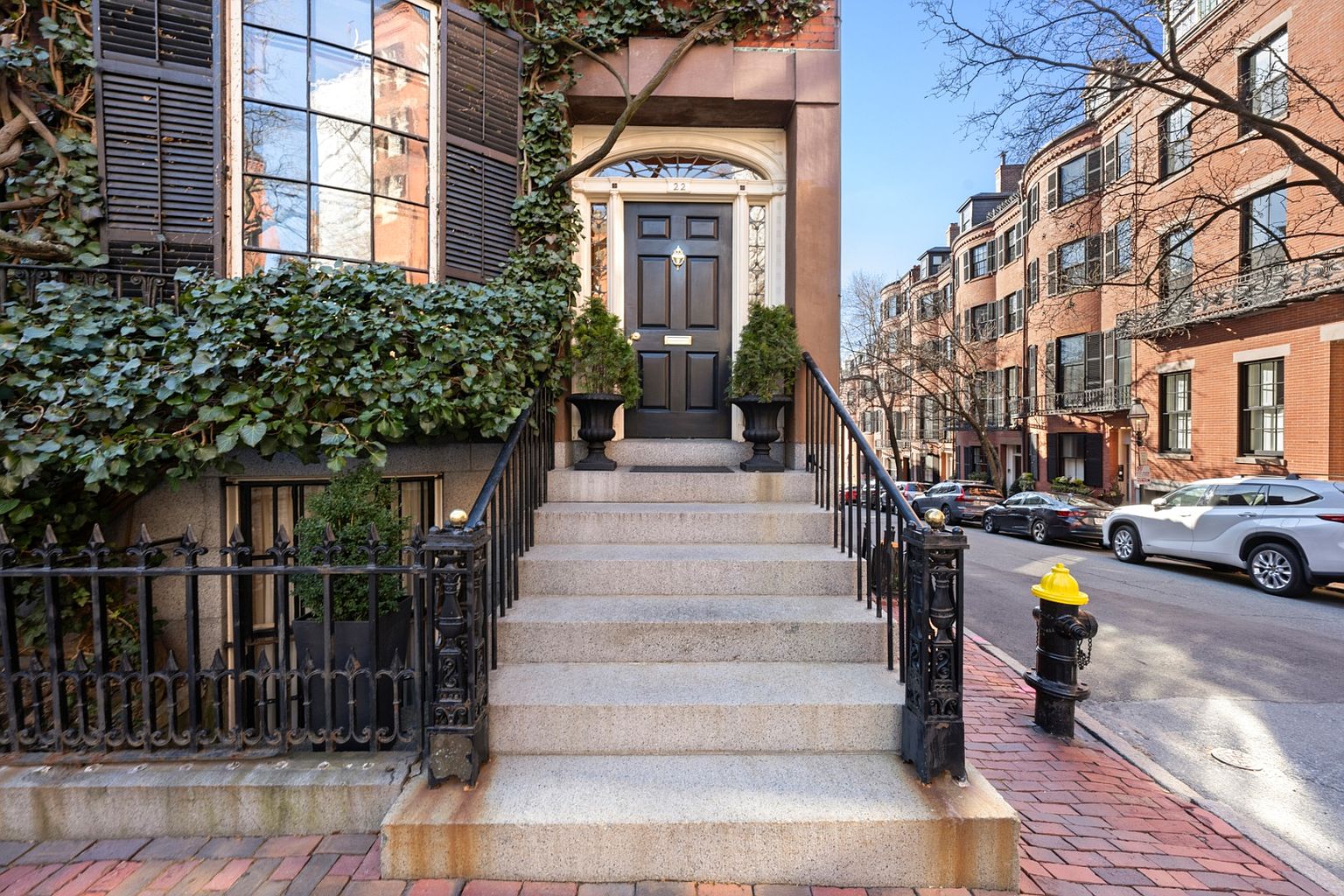 This image showcases the grand entryway of a brick townhouse, emphasizing its classic and elegant appeal. A set of stone steps leads up to a dark-colored front door, flanked by manicured greenery and ornate black iron railings. The facade is partially covered in ivy, adding a touch of old-world charm and highlighting the property's established character.