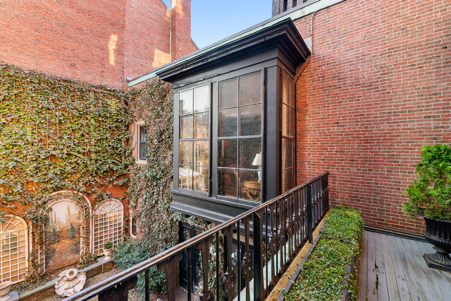 This image showcases a charming outdoor space, likely a patio or balcony, featuring a black railing and a view of a brick courtyard. The courtyard is adorned with lush greenery, including ivy-covered walls and potted plants, creating a serene and inviting atmosphere. A unique architectural feature, possibly a bay window or enclosed porch, adds character to the scene.