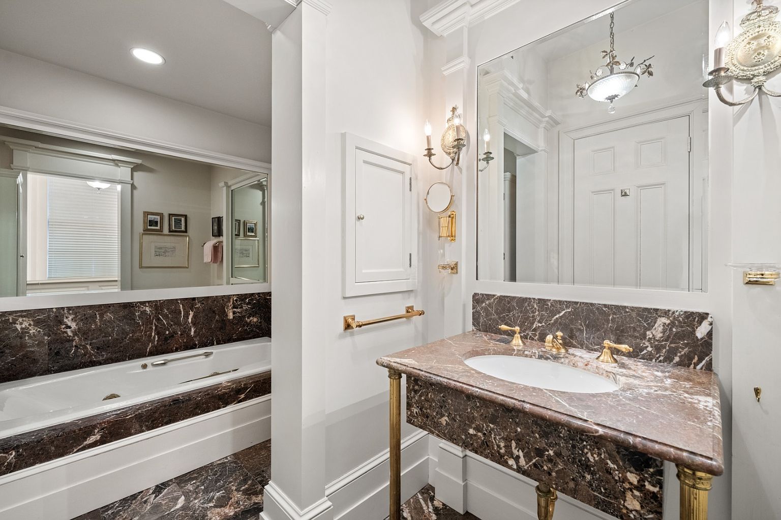 This is a luxurious primary bathroom featuring a marble countertop vanity with gold fixtures and a large mirror reflecting a doorway and chandelier. A soaking tub with marble surround is visible to the left, complemented by matching marble flooring. The bathroom exudes elegance and sophistication with its classic design elements.