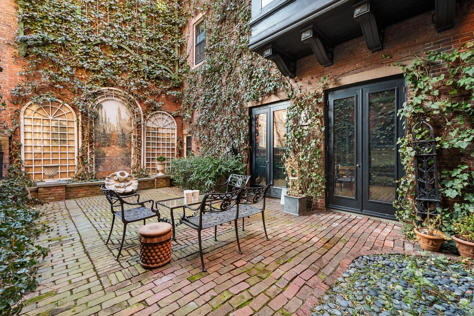 This image showcases a charming courtyard garden, featuring brick paving and walls covered in lush ivy. A wrought-iron seating area with a glass-topped table is centrally located, complemented by decorative architectural elements like arched window frames and a painted mural. The overall impression is one of a secluded, tranquil outdoor space, perfect for relaxation and entertaining.